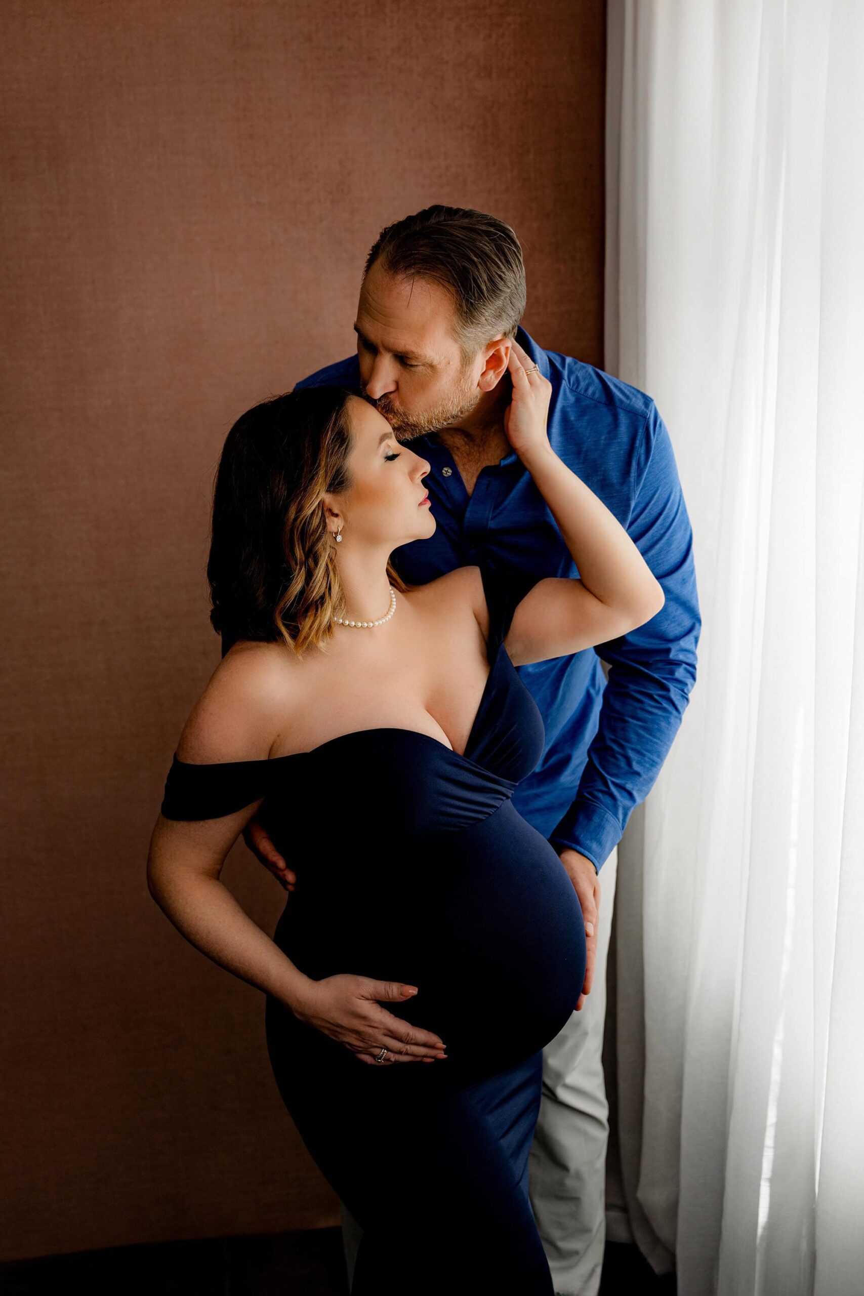 expectant couple poses in waxhaw, nc studio with mom wearing a navy maternity client closet dress and pearls and dad wearing complimentary blue while kissing mom's forehead