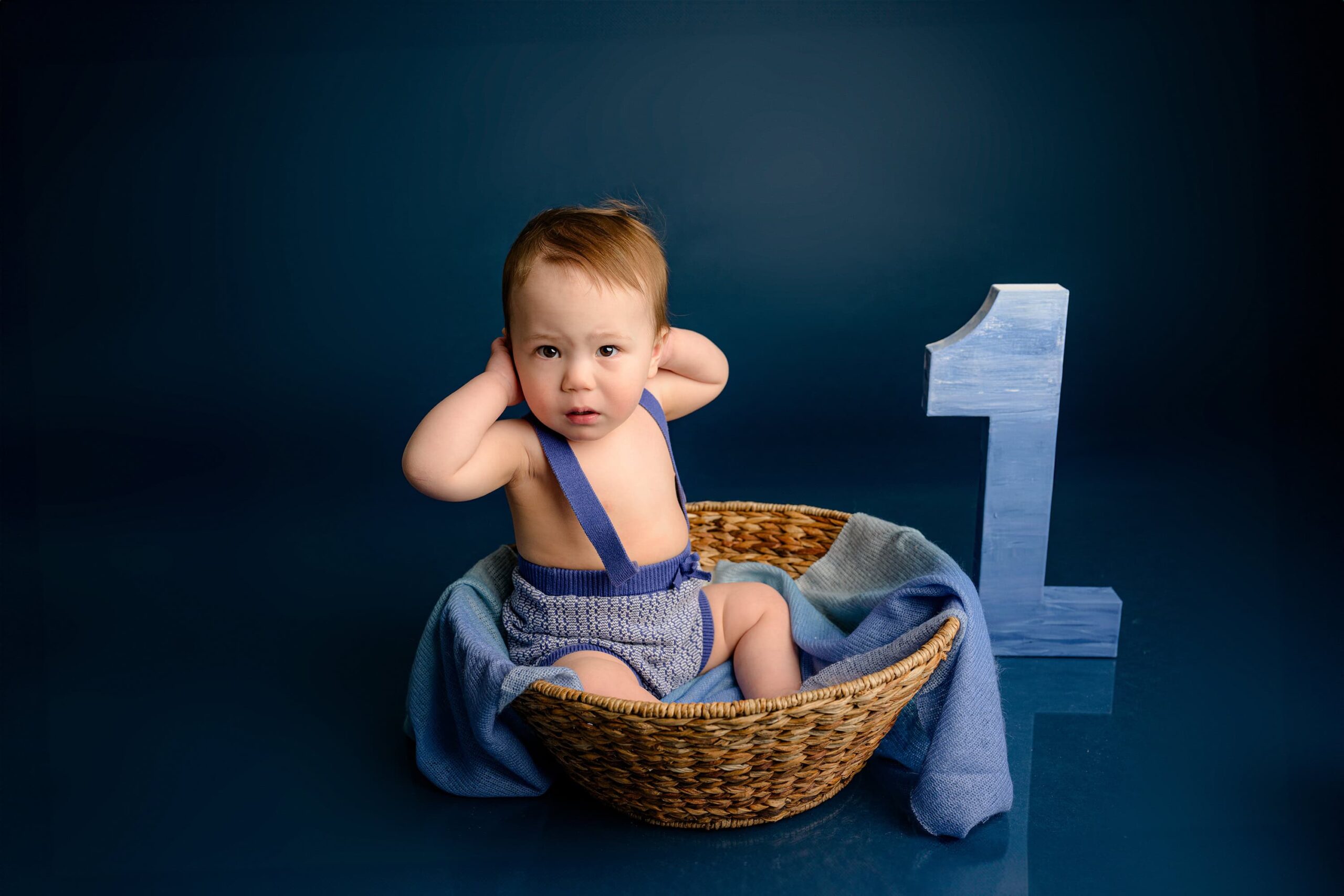 A one-year-old baby boy sits in a basket with a multicolored blue wrap under him, hands up by his head, with a large number one beside him on a blue studio background.