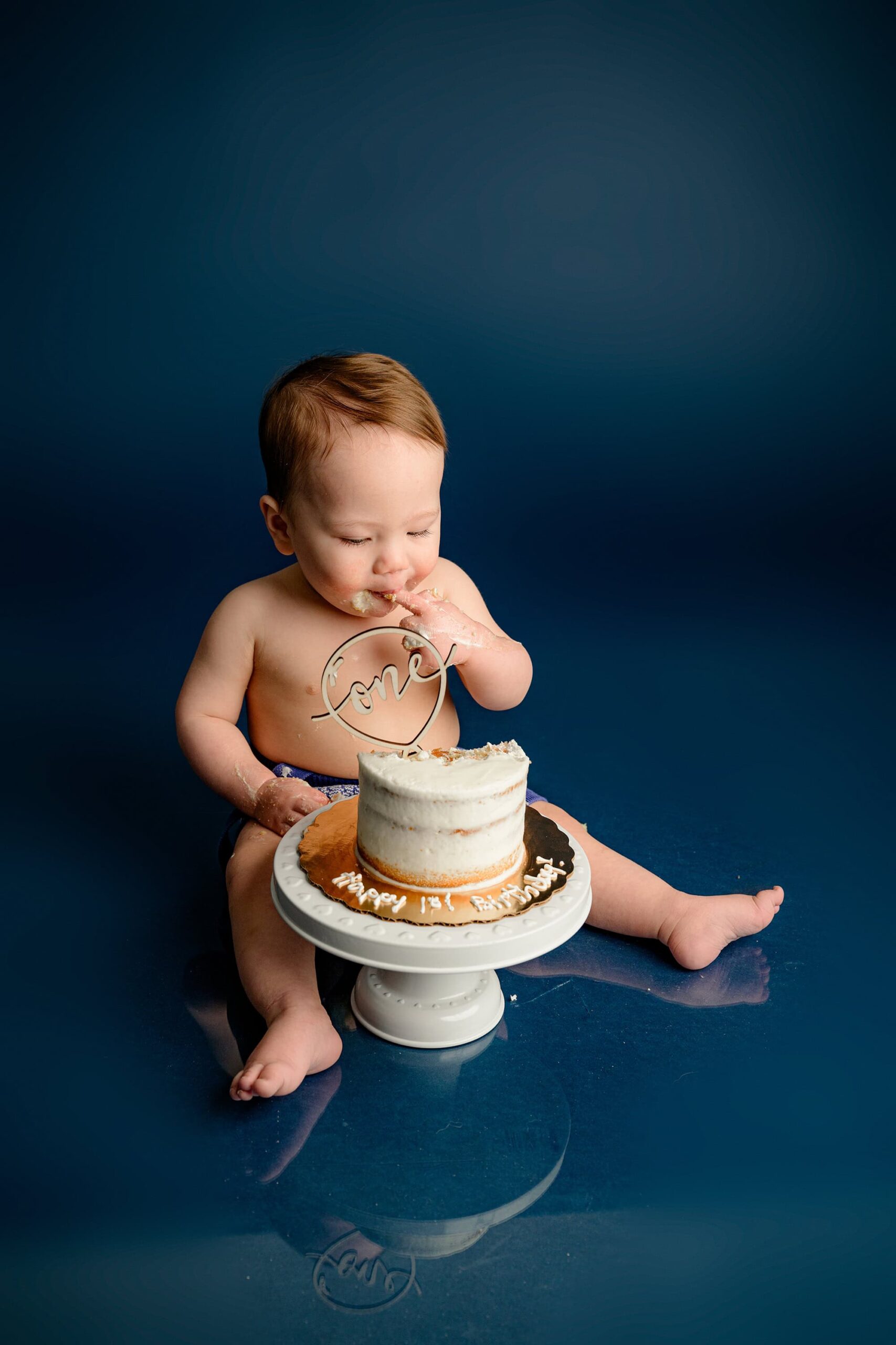A one-year-old baby boy carefully takes a small taste of his smash cake while looking down at it, savoring every bite during his birthday photoshoot on a blue background.