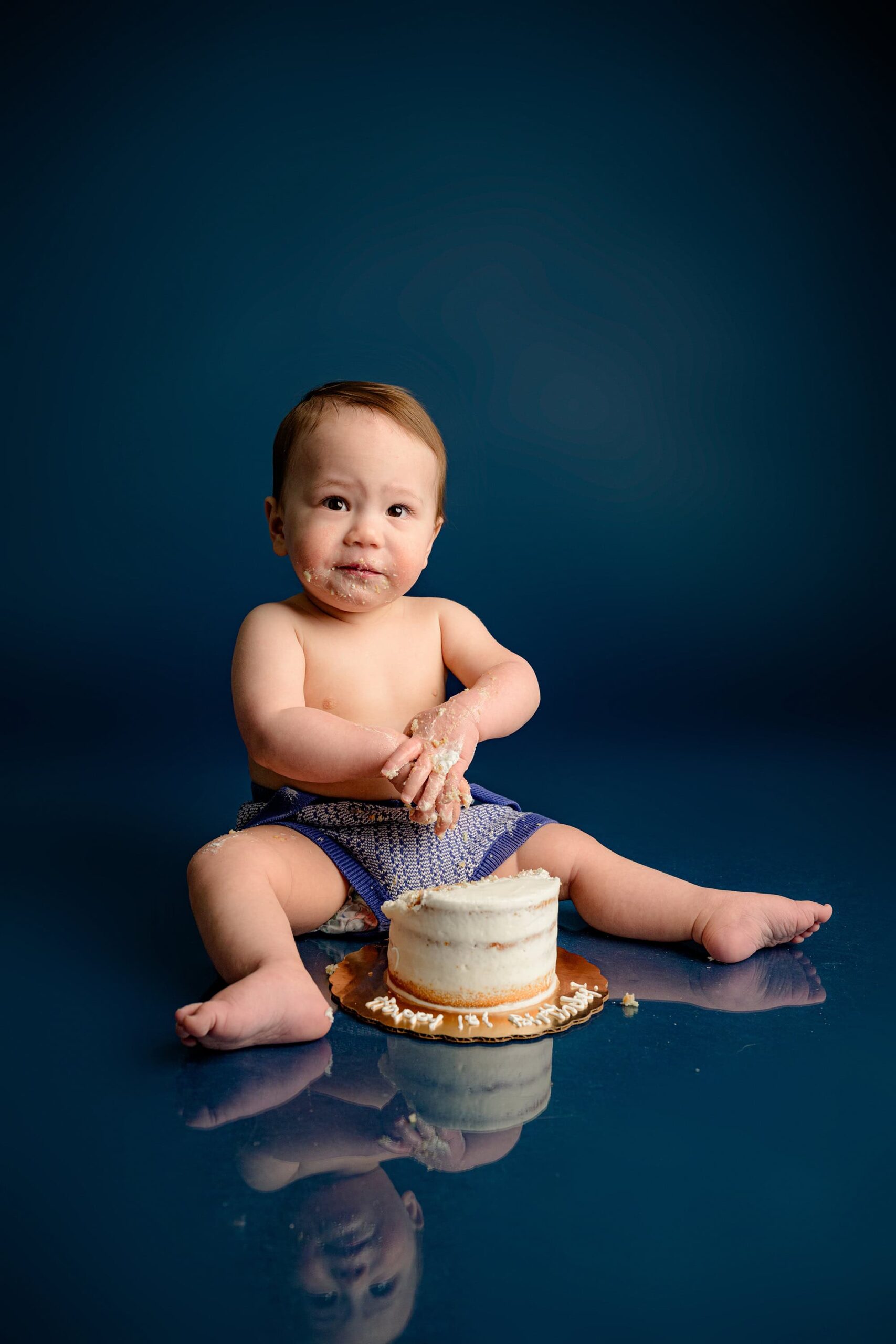 A one-year-old baby boy plays with his cake-covered hands, enjoying the messy fun of his birthday cake smash session on a blue studio background.