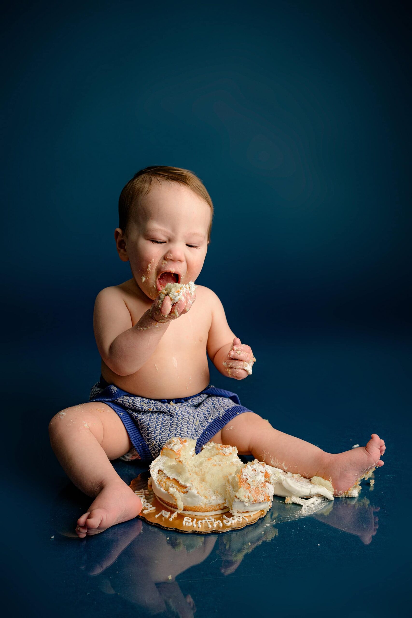 A one-year-old baby boy looks down at his smash cake, taking a huge bite, fully immersed in the joy of his first birthday celebration on a blue background.