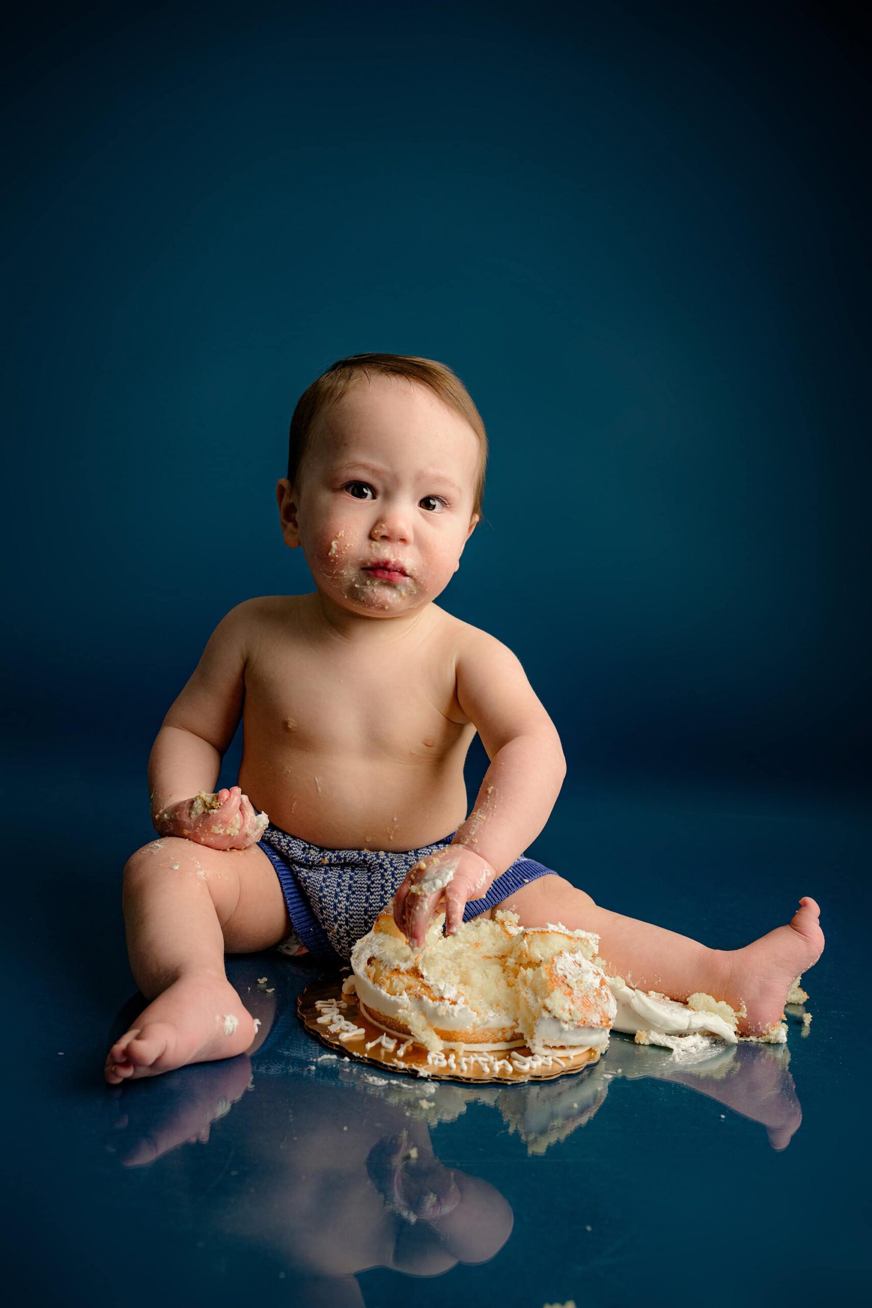 A one-year-old baby boy excitedly smashes his multilayered, naked-iced cake, which has fallen off the cake plate, during his birthday photoshoot on a blue background.