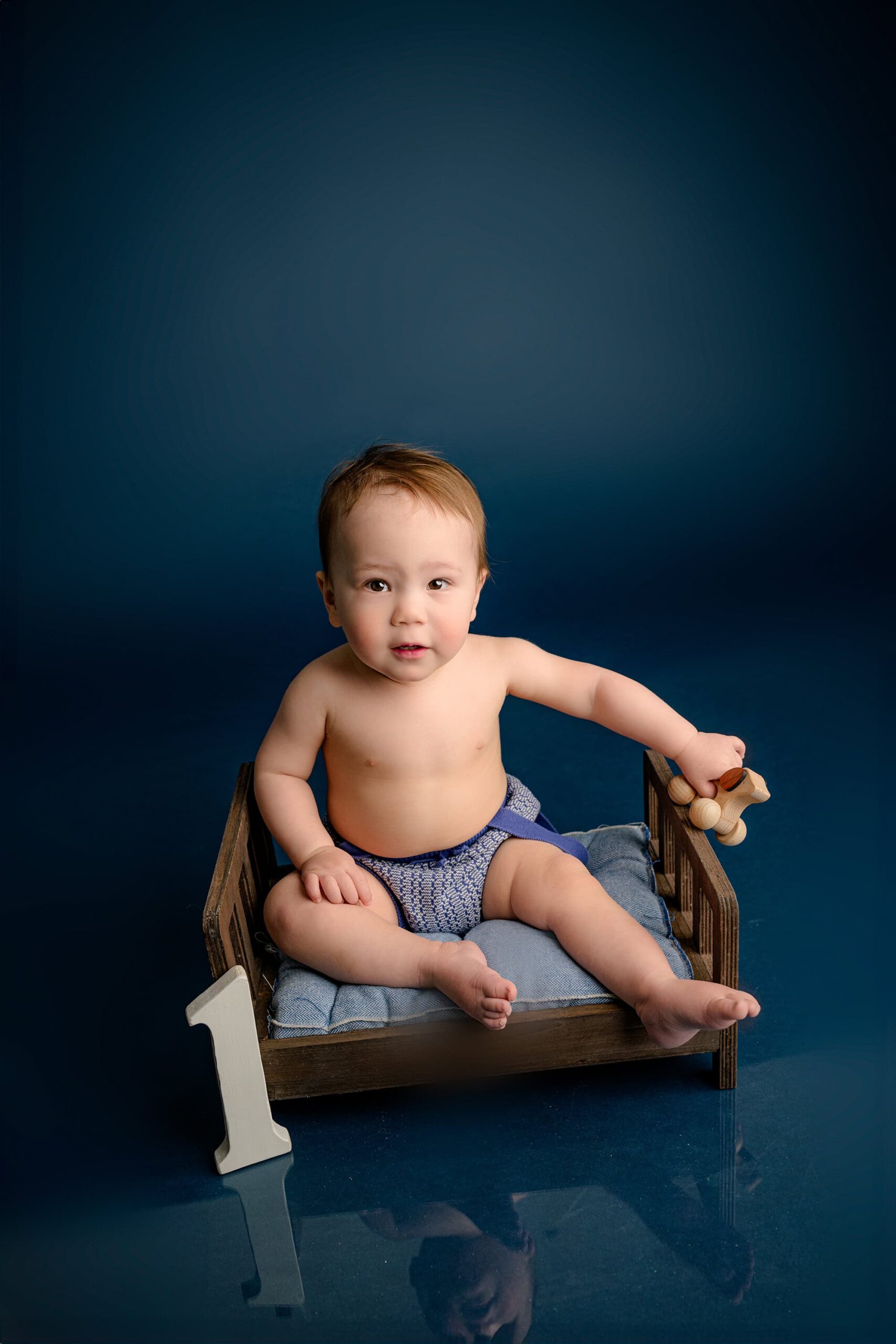 A one-year-old baby boy in a cream and blue knit outfit sits on a brown bed with a small number one beside him, smiling and playing with a toy on a blue studio background.