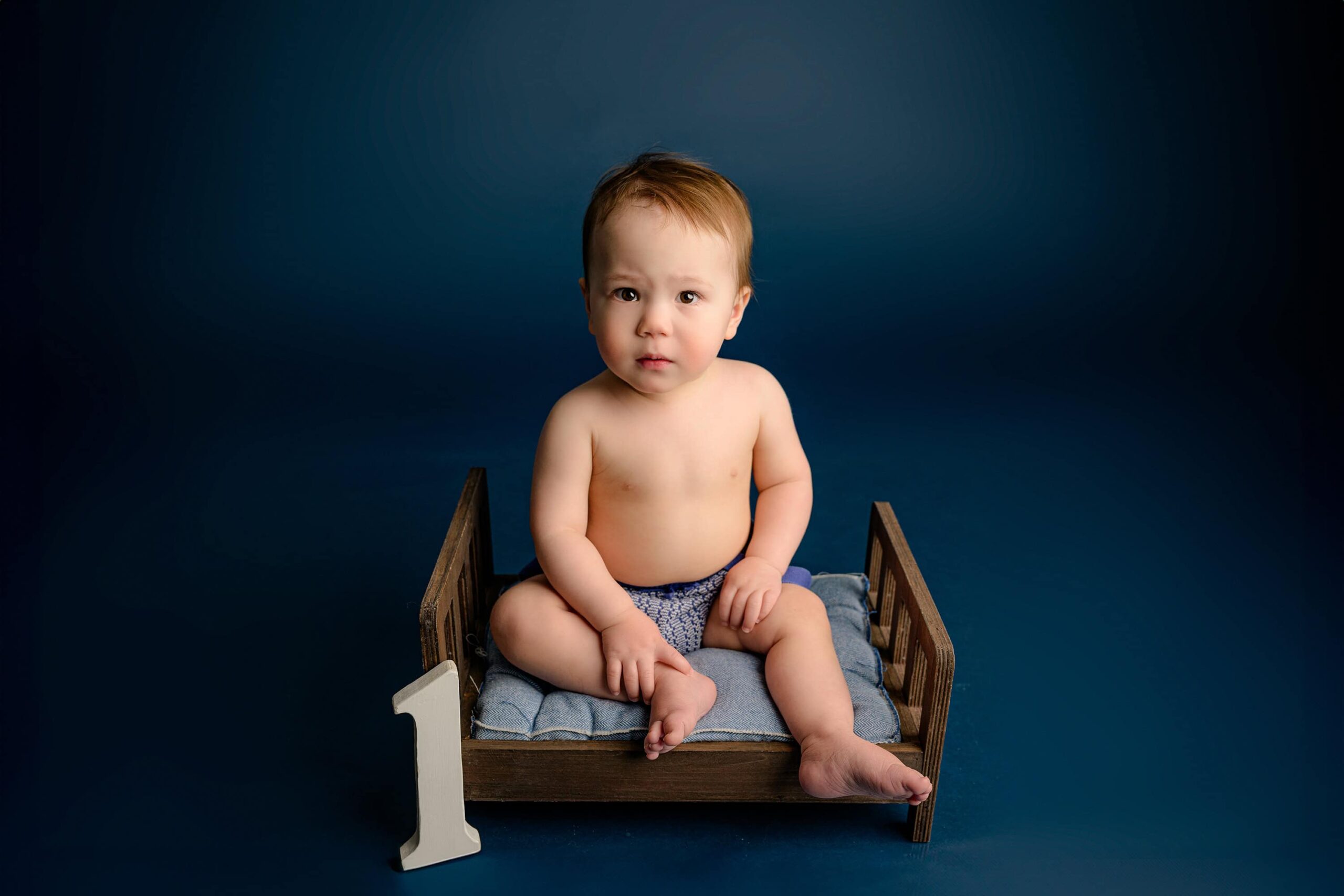 A one-year-old baby boy wearing a cream and blue knit outfit sits on a blue studio background, looking directly at the camera during his birthday photoshoot.