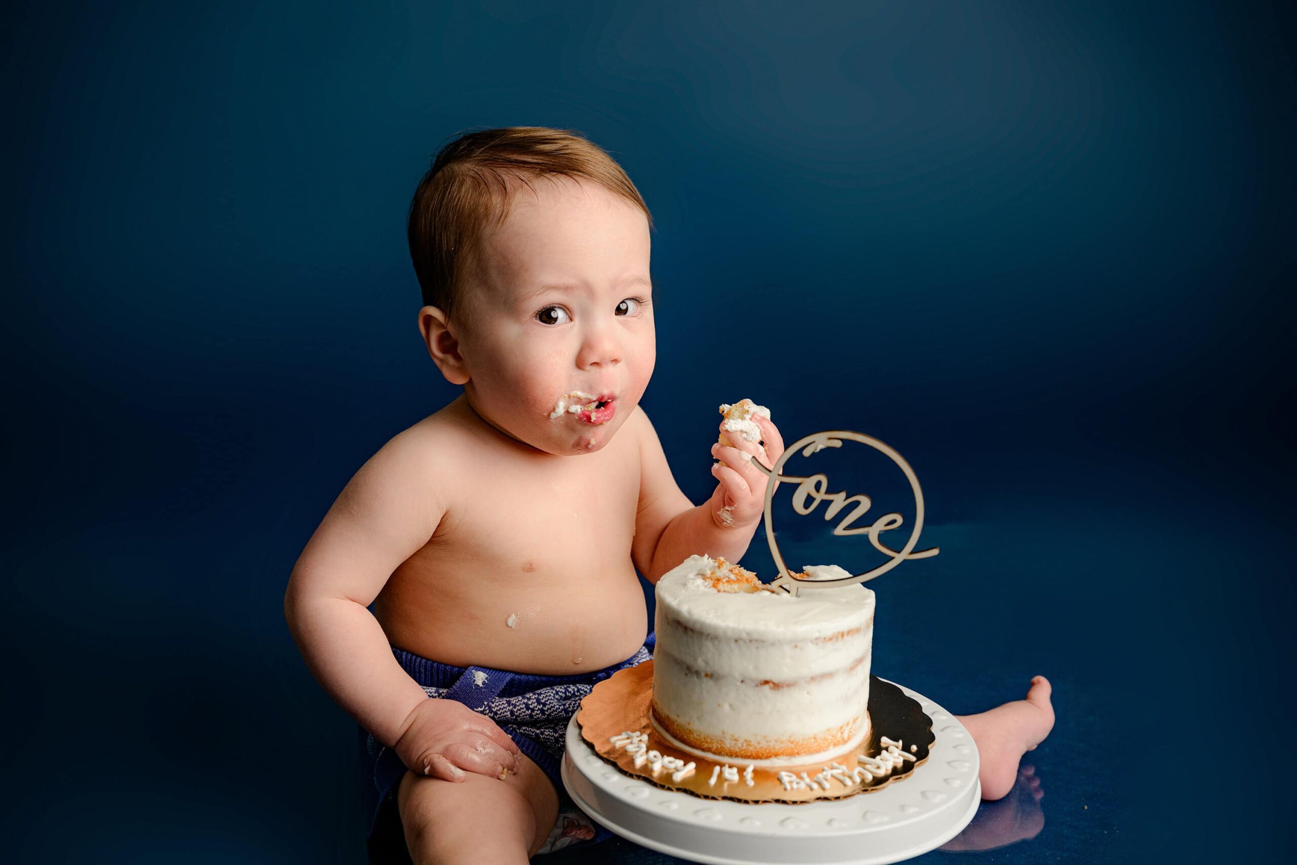 A one-year-old baby boy wearing a cream and blue knit outfit gives the best studio session side-eye while eating his smash cake during his birthday photoshoot on a blue background.