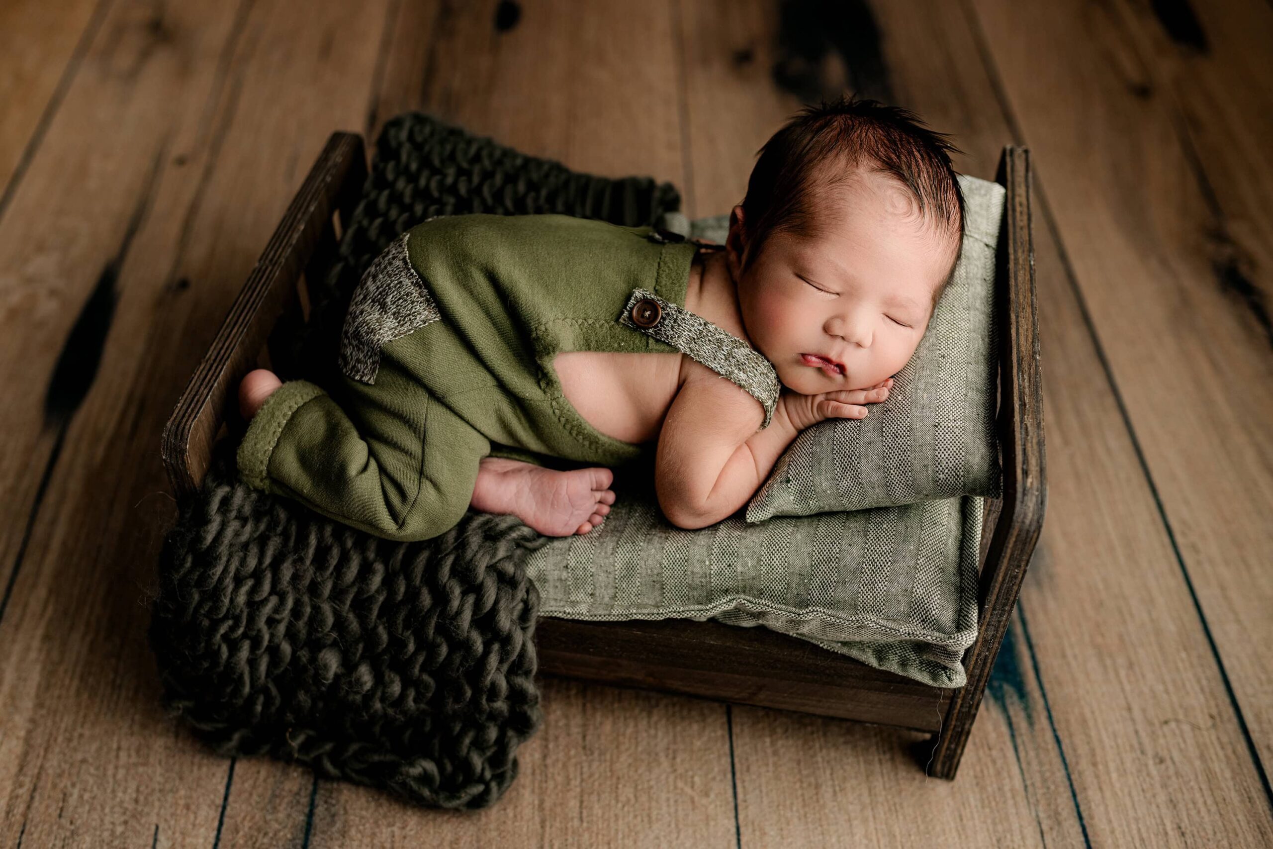 A charming image of a baby dressed in green overalls, posed on his side and resting on a pillow. The baby is surrounded by a warm wooden floor, a brown bed, and soft green bedding, creating a cozy and peaceful atmosphere in this adorable setting.
