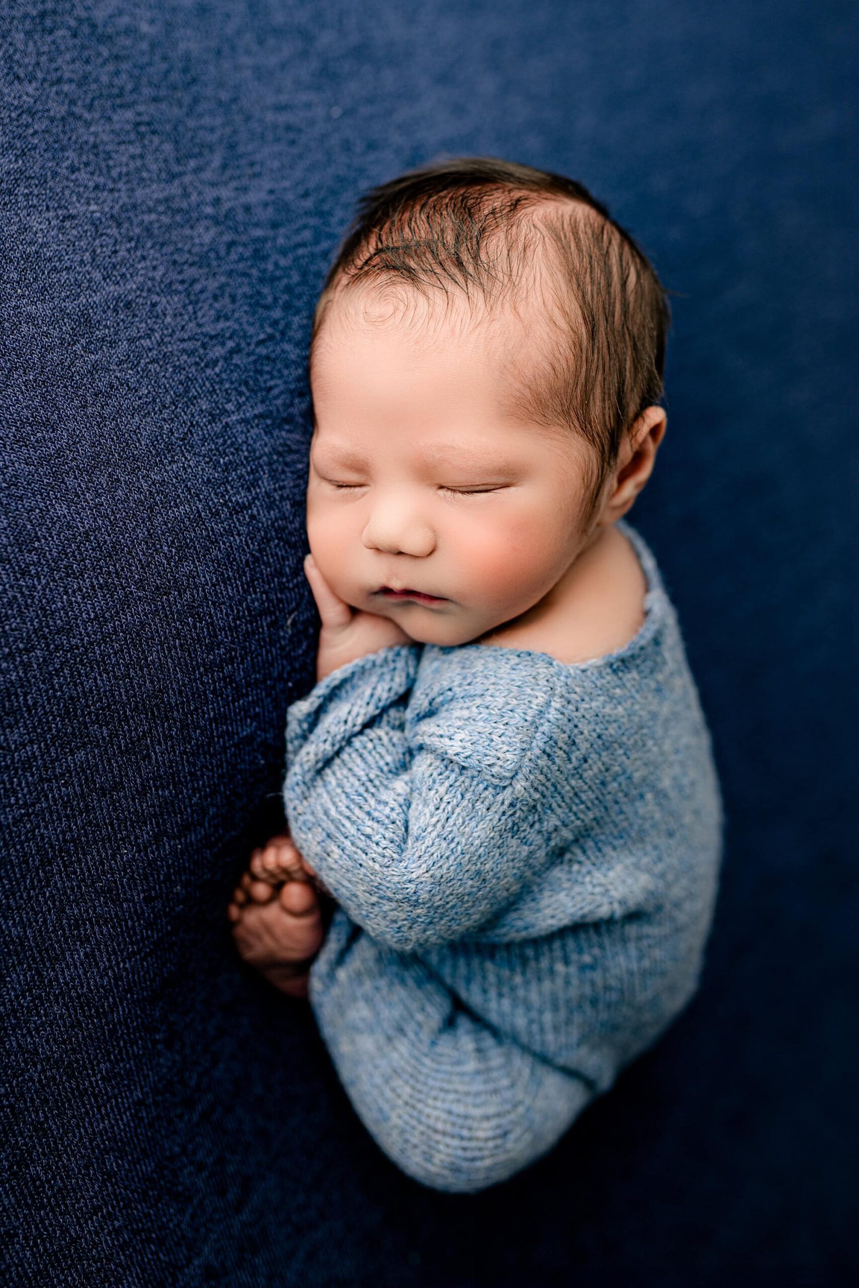 A newborn baby boy in a blue knit outfit is posed on a soft blue background, resting with one hand under his cheek and his tiny feet curled under him.