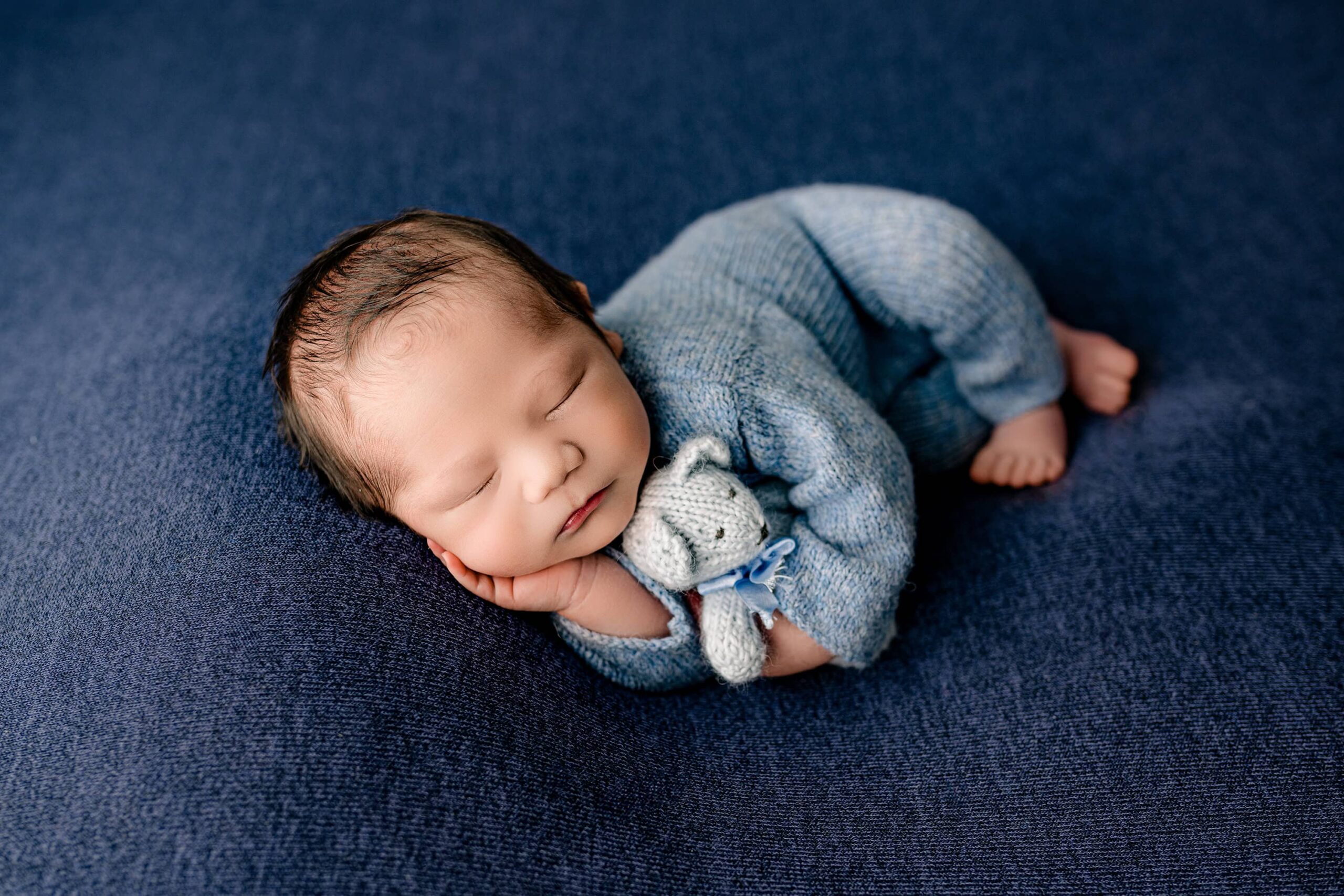 A newborn baby boy wears a blue knit outfit and is posed on a soft blue background, peacefully resting with one hand under his chin and a small teddy bear beside him.