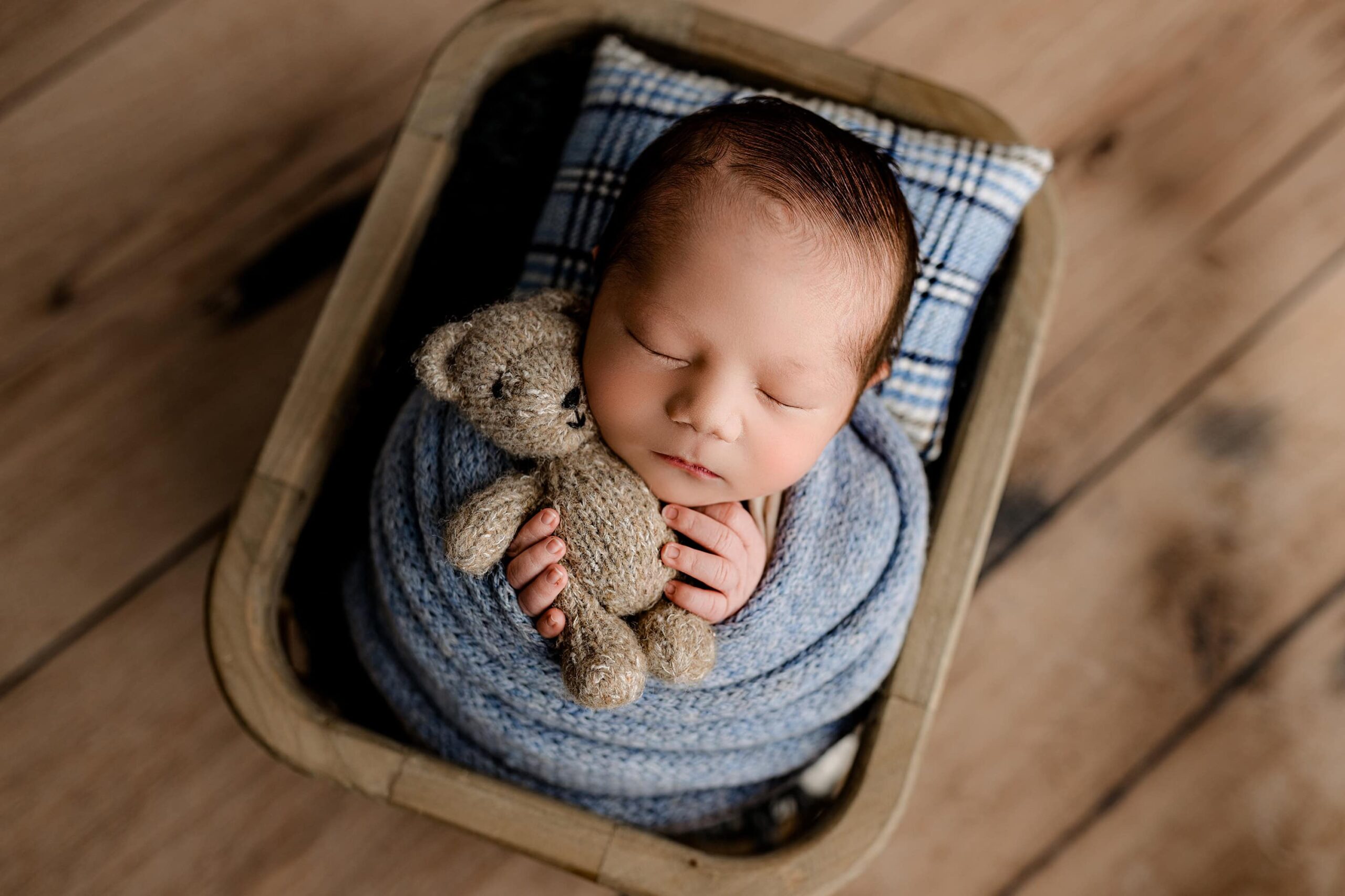 A peaceful newborn session photo of a baby wrapped in a soft blue fabric, nestled comfortably in a cozy basket on a wooden floor. The baby gently snuggles a brown teddy bear, radiating warmth and tranquility in this adorable moment.