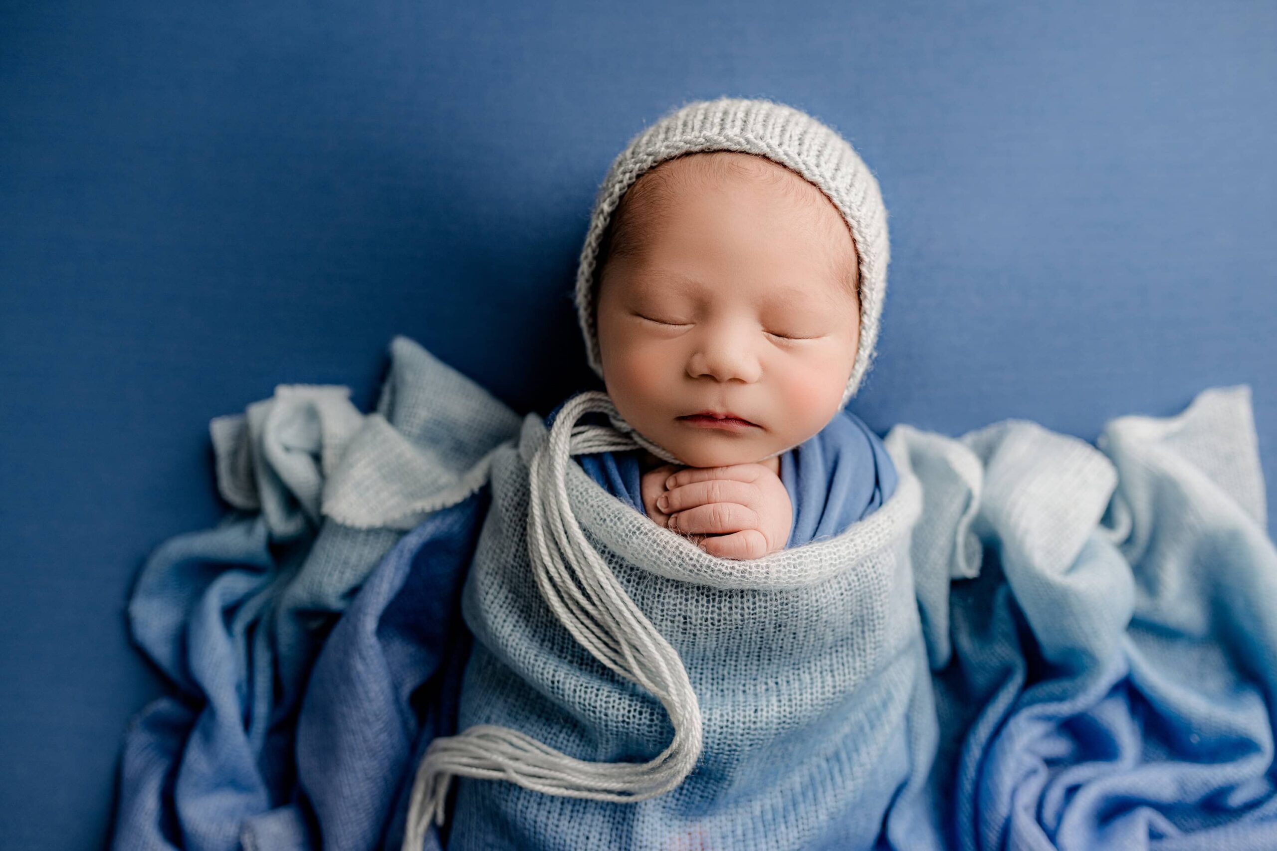 An adorable newborn photo featuring a baby posed on a soft blue background, wearing a matching blue bonnet. The baby is wrapped in a colorful, multicolored knit wrap, creating a cozy and vibrant look in this sweet moment.