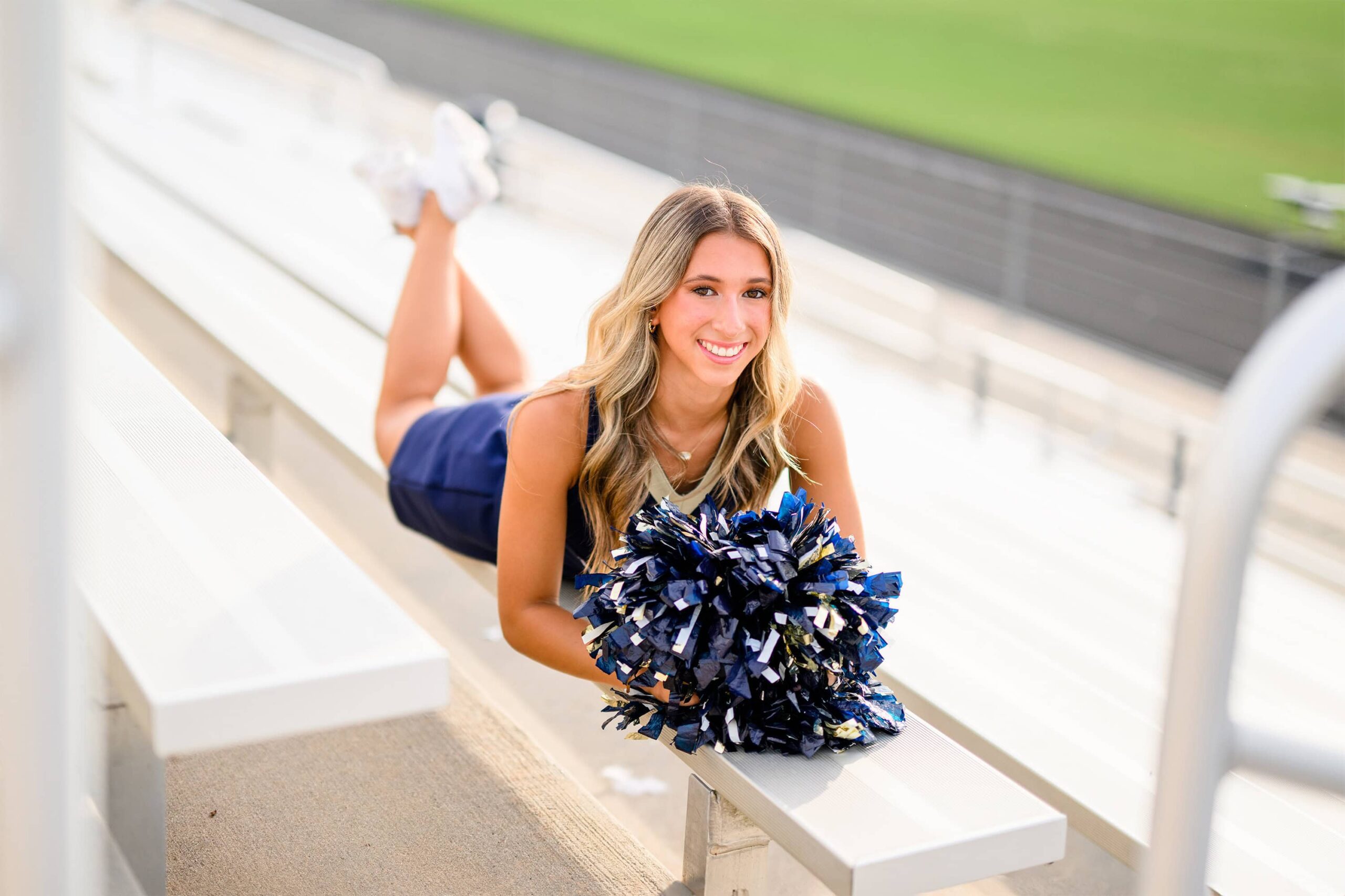 A blonde cheerleader from Cuthbertson High School in Waxhaw, NC, lies on her stomach on the bleachers, smiling with her poms placed in front of her.
