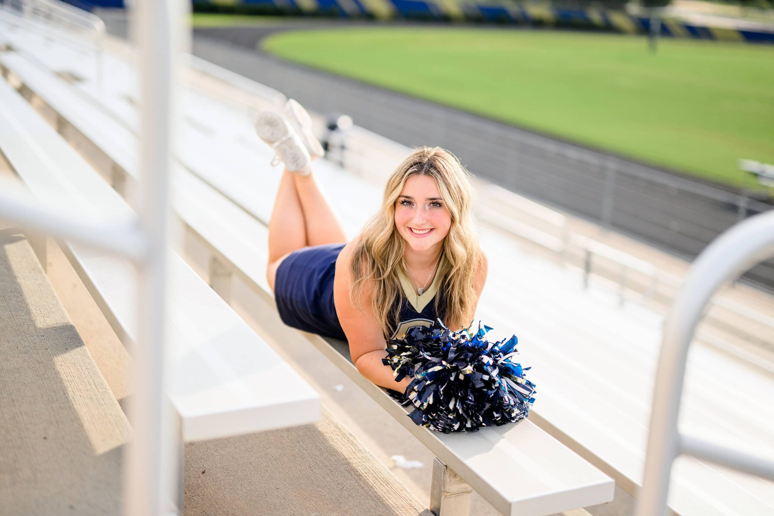 Olivia, a senior cheerleader with blonde hair from Cuthbertson High School, poses on her stomach on the bleachers, her poms resting in front of her as she smiles.