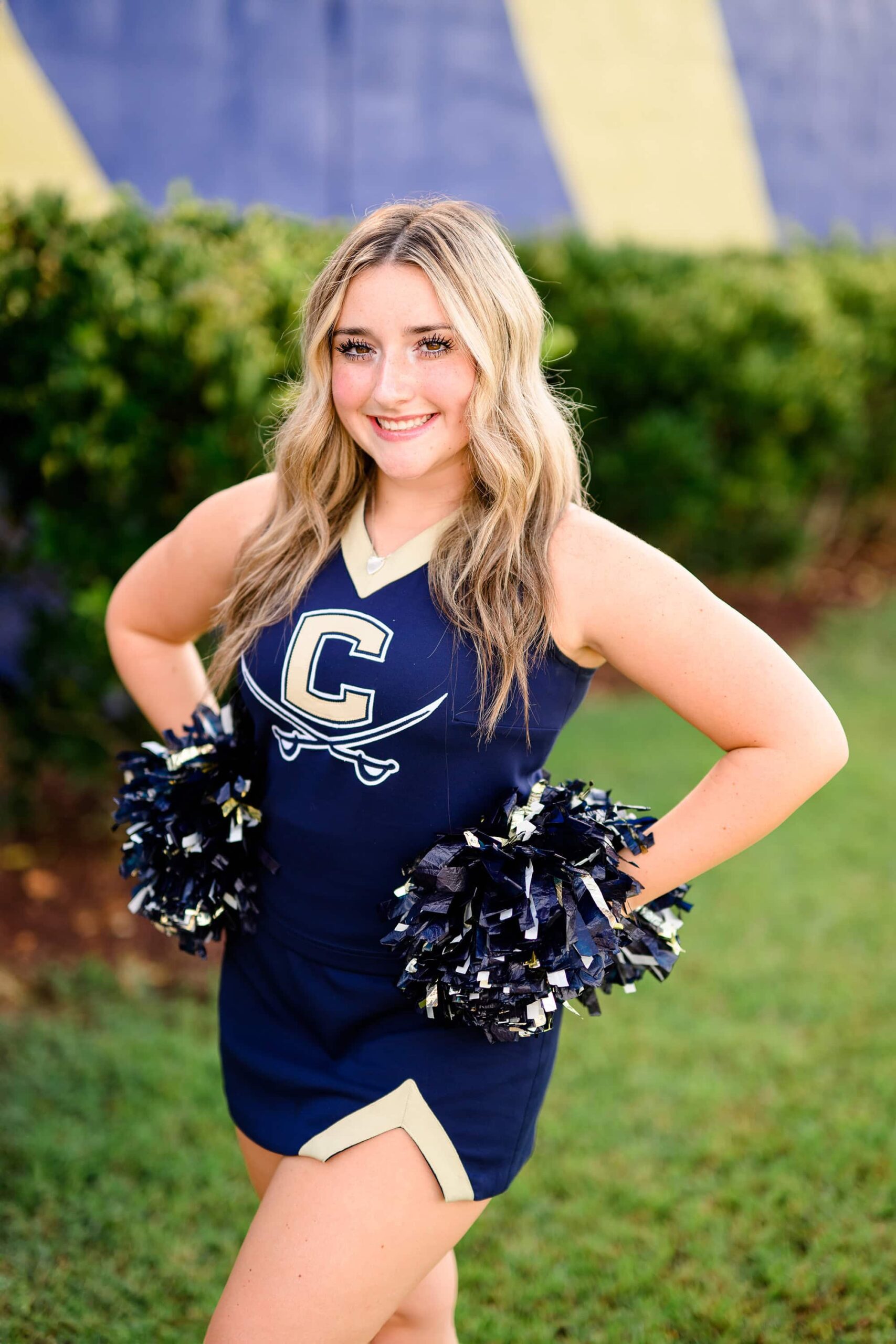 Olivia, a cheerleader from Cuthbertson High School, stands confidently with her hands on her hips, holding poms, and smiling.