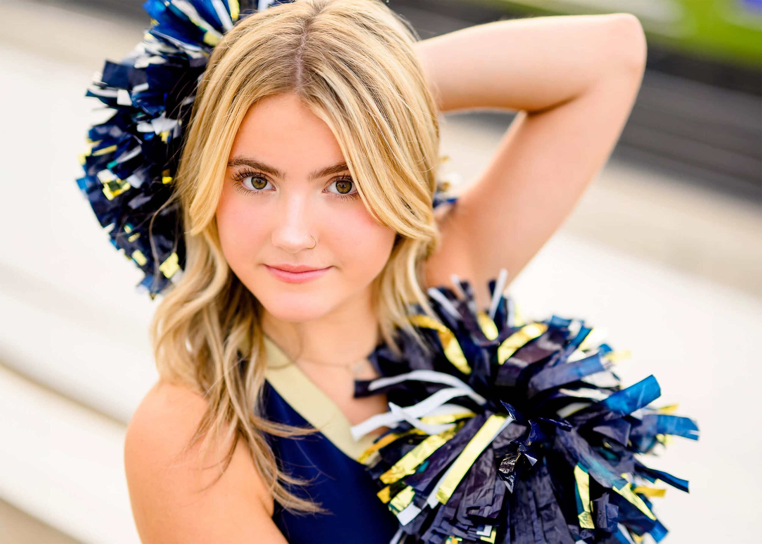A blonde cheerleader from Cuthbertson High School sits on the bleachers with her poms placed in front of and behind her, smiling brightly during her senior cheer photos.