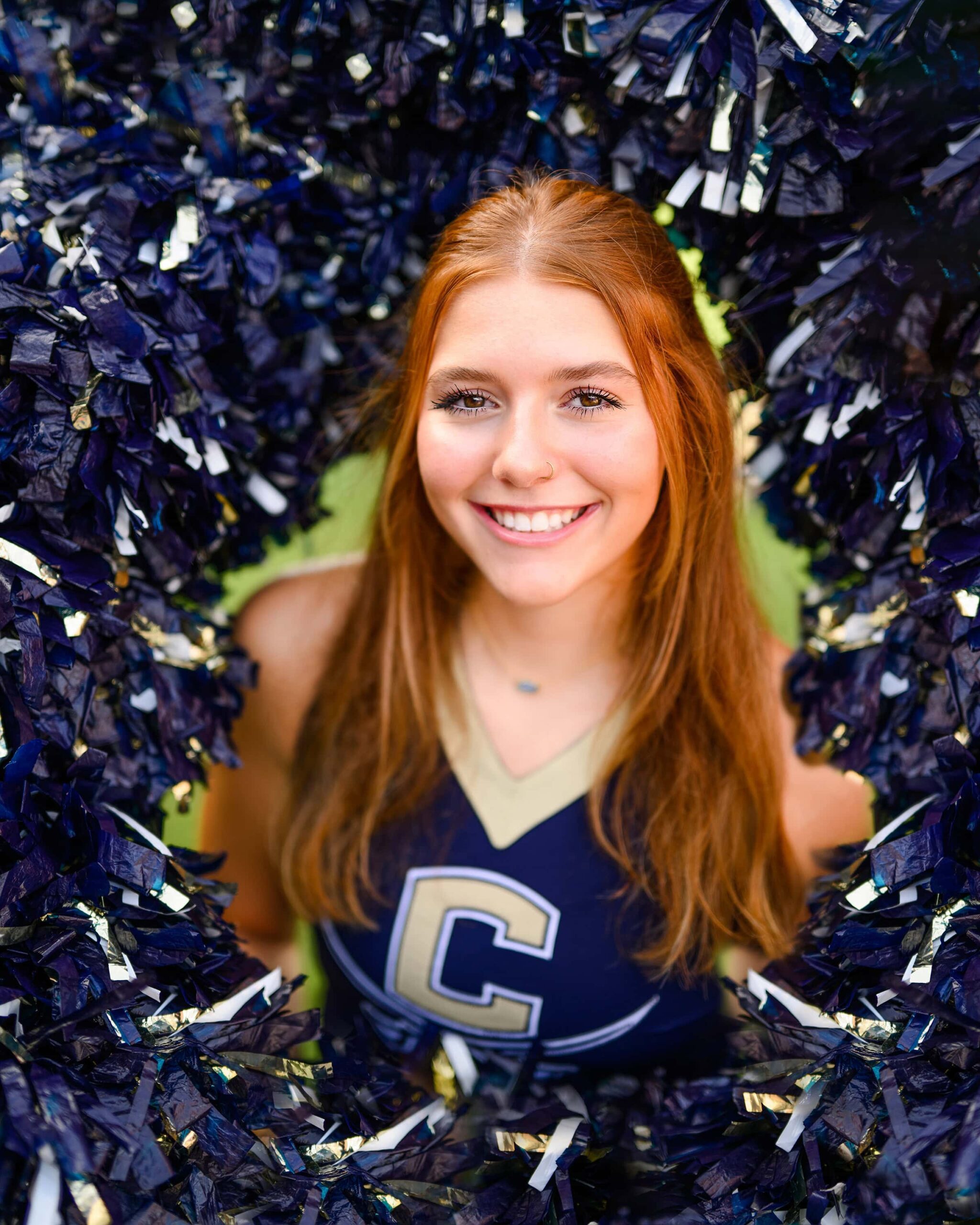 A red-haired cheerleader from Cuthbertson High School in the center of a pom-pom ring, framed by a circle of poms around her head, looking up at the camera with a bright expression.