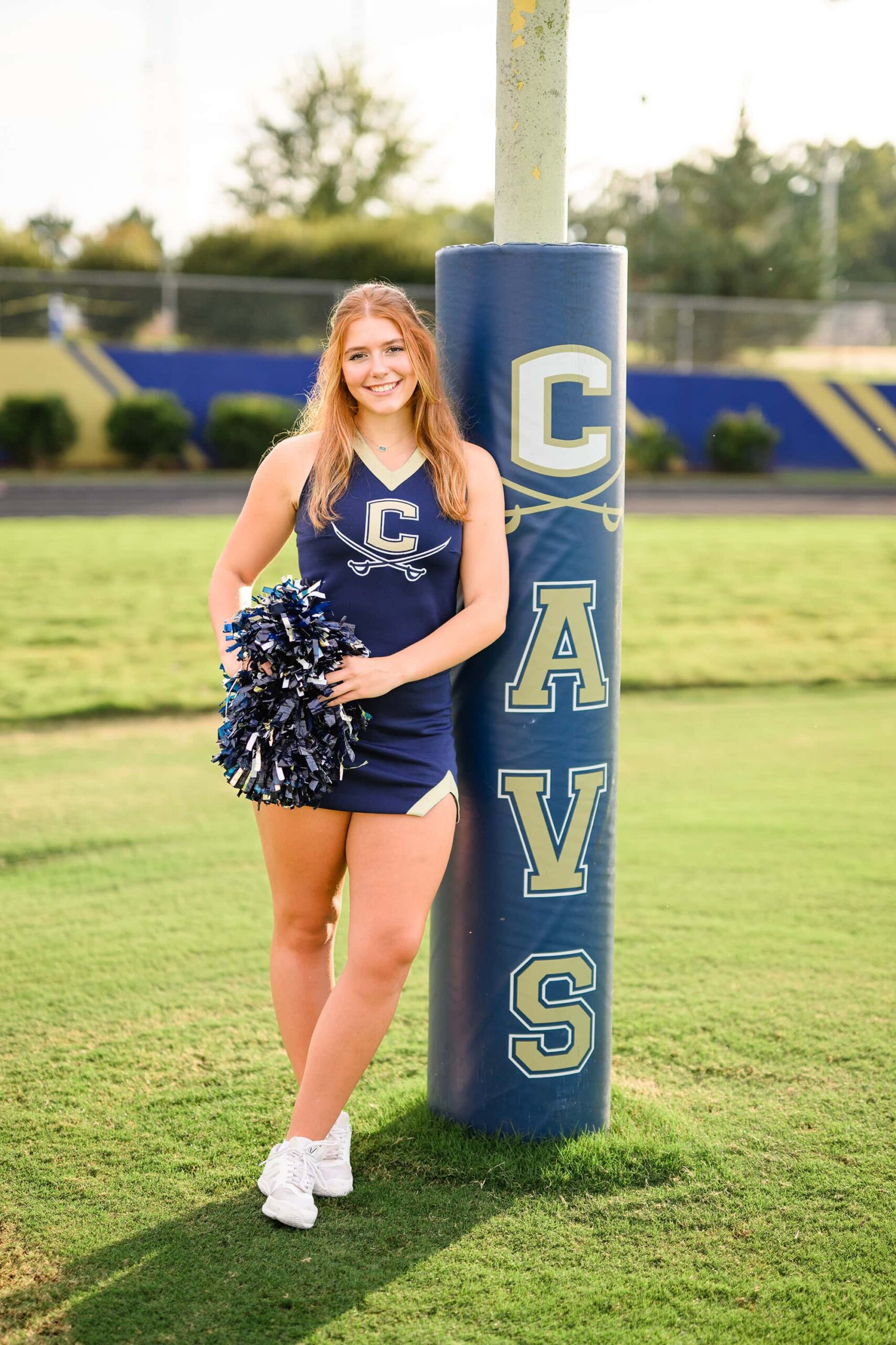 A full-body image of a redheaded cheerleader from Cuthbertson High School in her uniform, leaning against a CAVS goalpost with a bright smile.