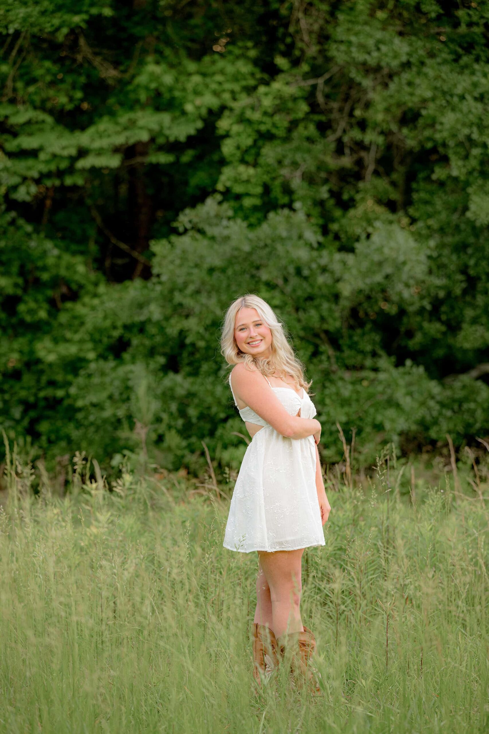 Kait poses in the field with her arm gently crossed over her waist, looking directly at the camera. Her feet are crossed, showcasing her cowboy boots beneath her flowing white dress.