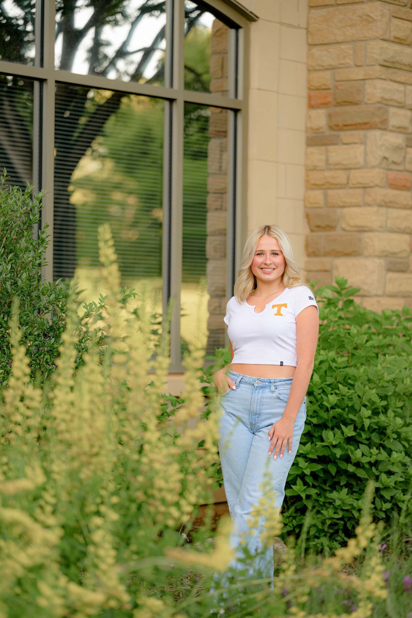 Amongst vibrant spring flowers, Kait poses with one hand casually in her jeans pocket while the other arm hangs down. Her legs are crossed as she enjoys this special moment as a Marvin Ridge High School senior.