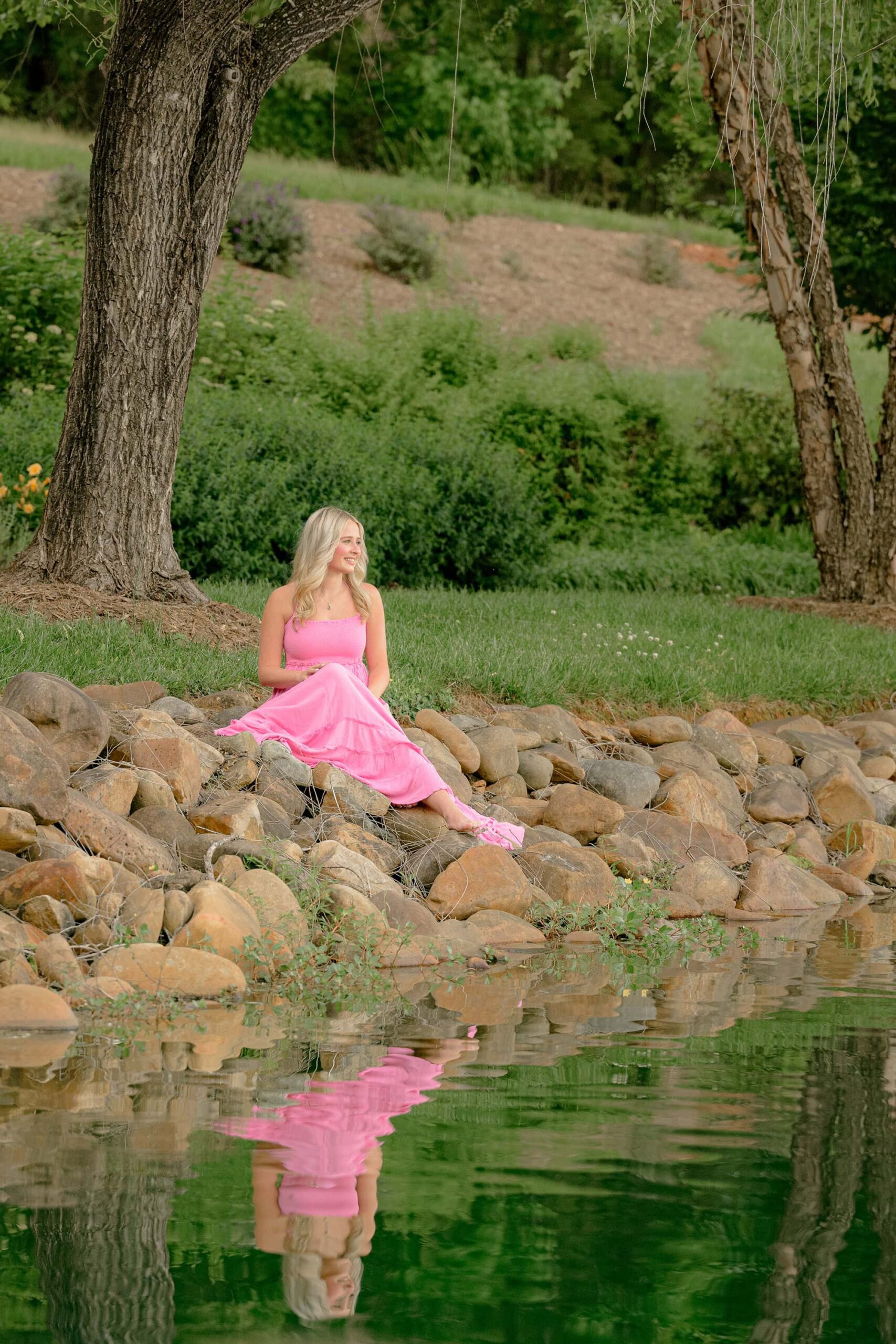 Seated on rocks beside the water, Kait gazes off to her left as the peaceful reflection shimmers in the water, capturing a serene Charlotte, NC, spring senior photo.