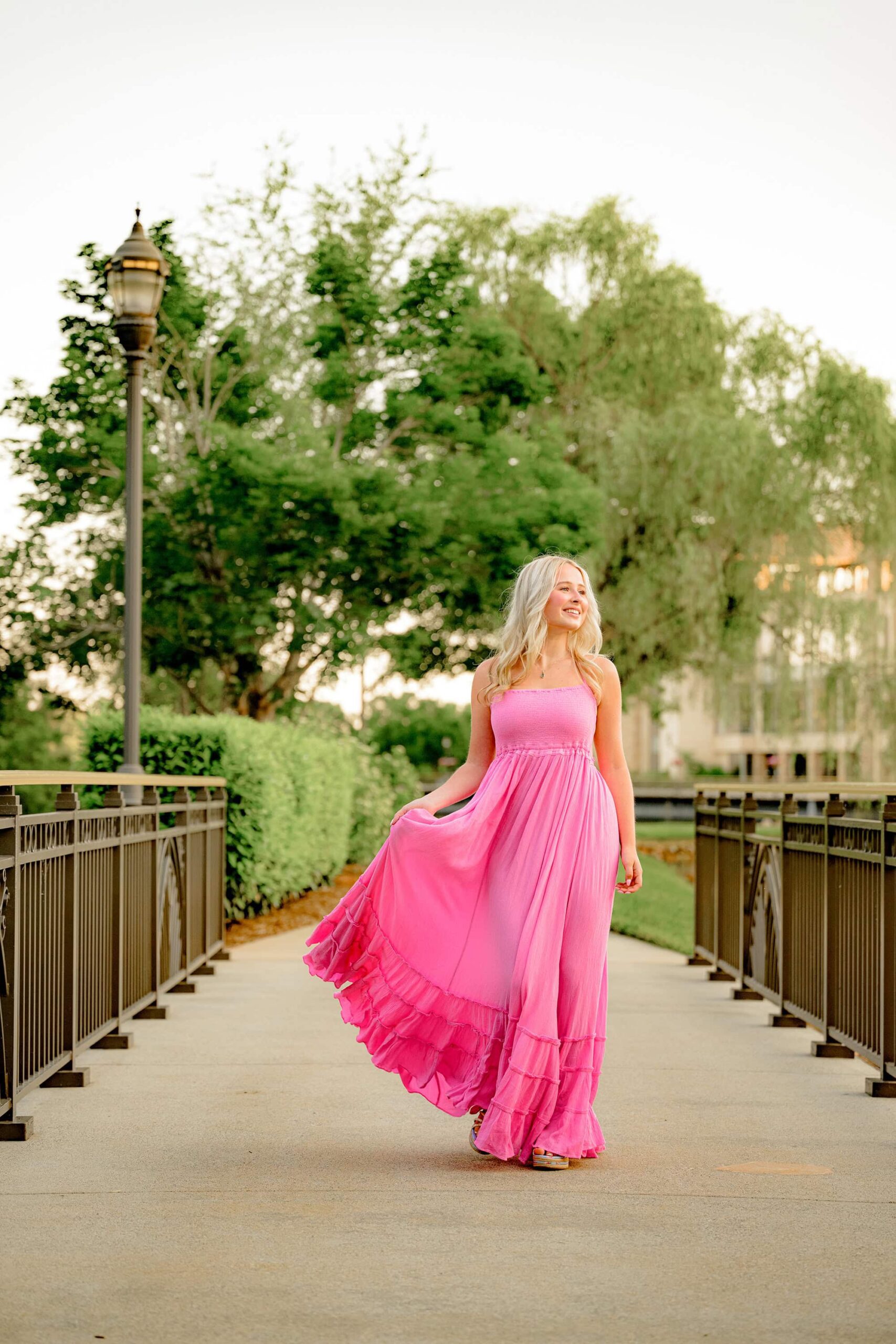 Kait walks gracefully across a small flat bridge, holding her dress as it blows in the wind. She looks off-camera, embracing the beauty of a spring senior photo in Charlotte, NC.