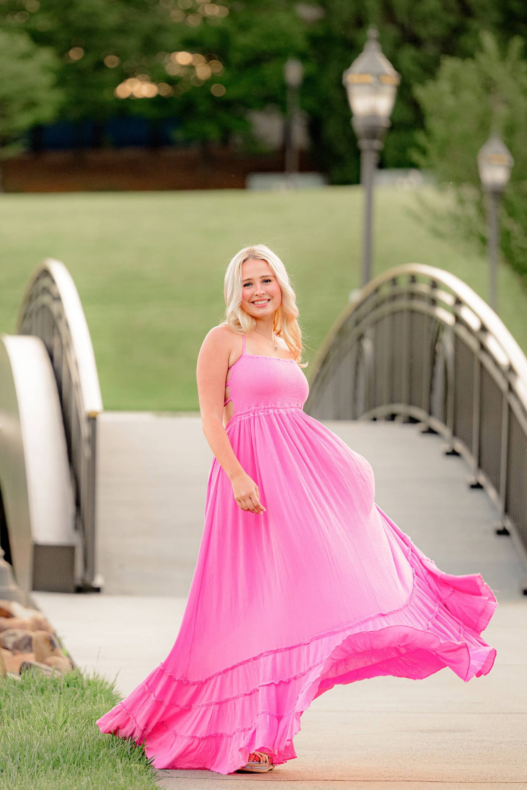 Kait, a Marvin Ridge High School senior, stands in front of an arched bridge, tossing her long pink flowy dress to create movement while smiling at the camera.