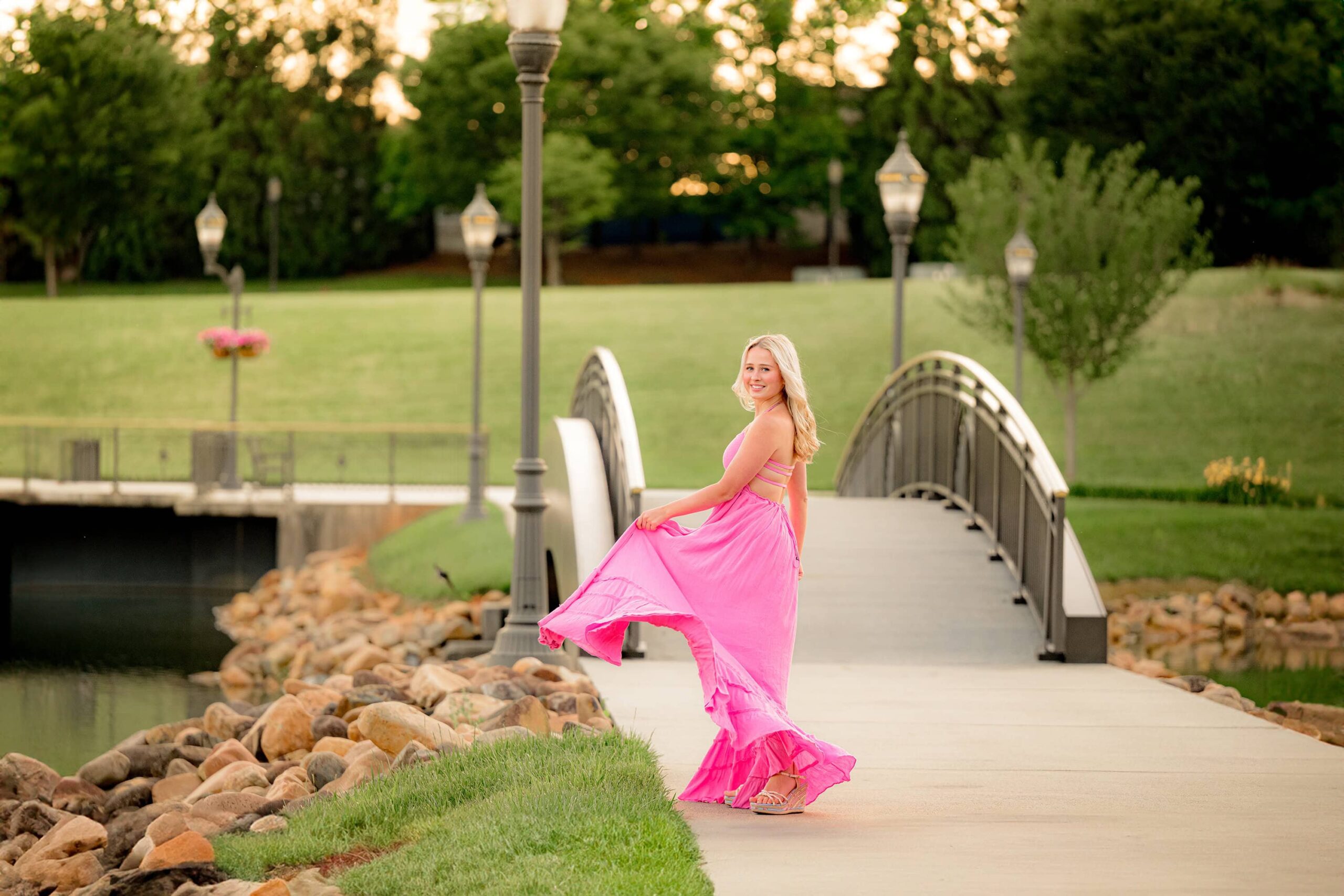 Walking away from the camera, Kait swishes her pink dress with an elegant twirl, glancing back over her shoulder. An arched bridge in the background adds to the dreamy Indian Land, SC setting.