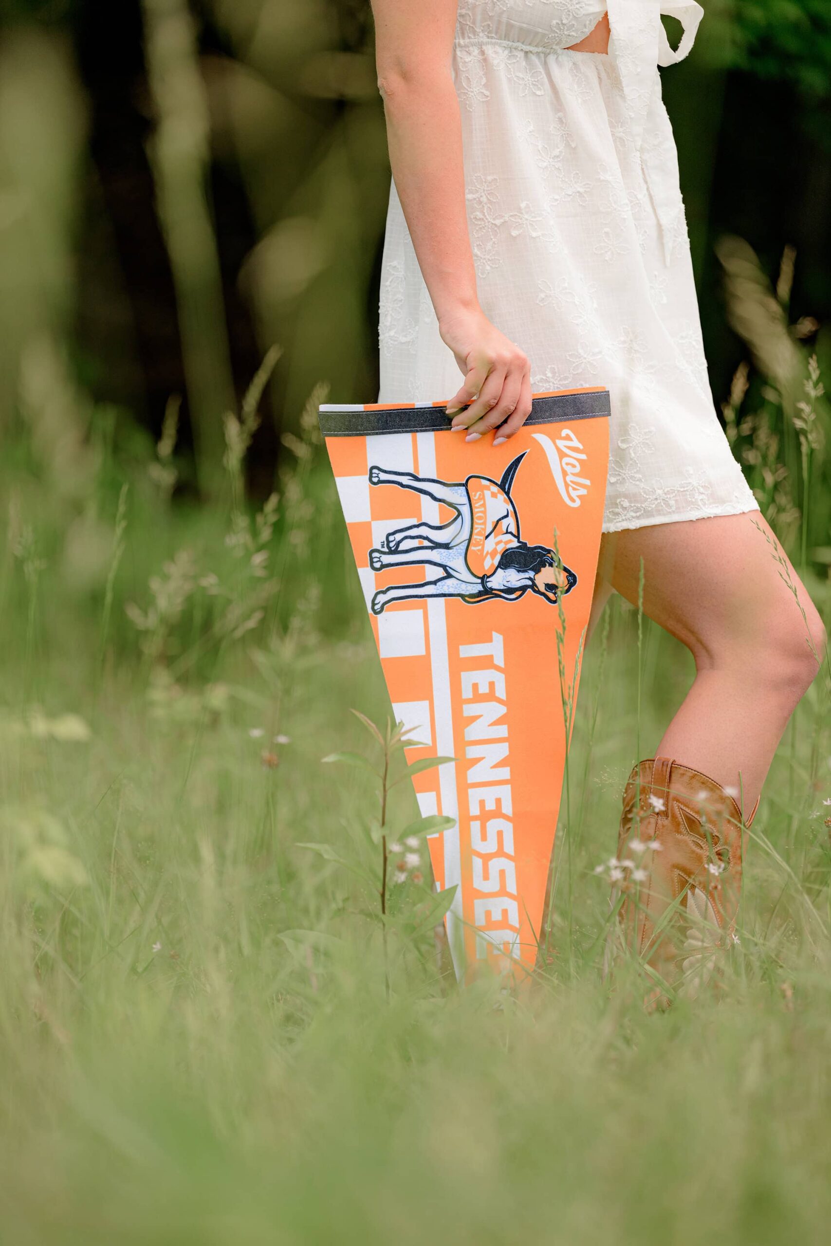 A close-up shot of Kait’s legs and white dress as her arm hangs down, drawing attention to a University of Tennessee pennant resting in the grass.