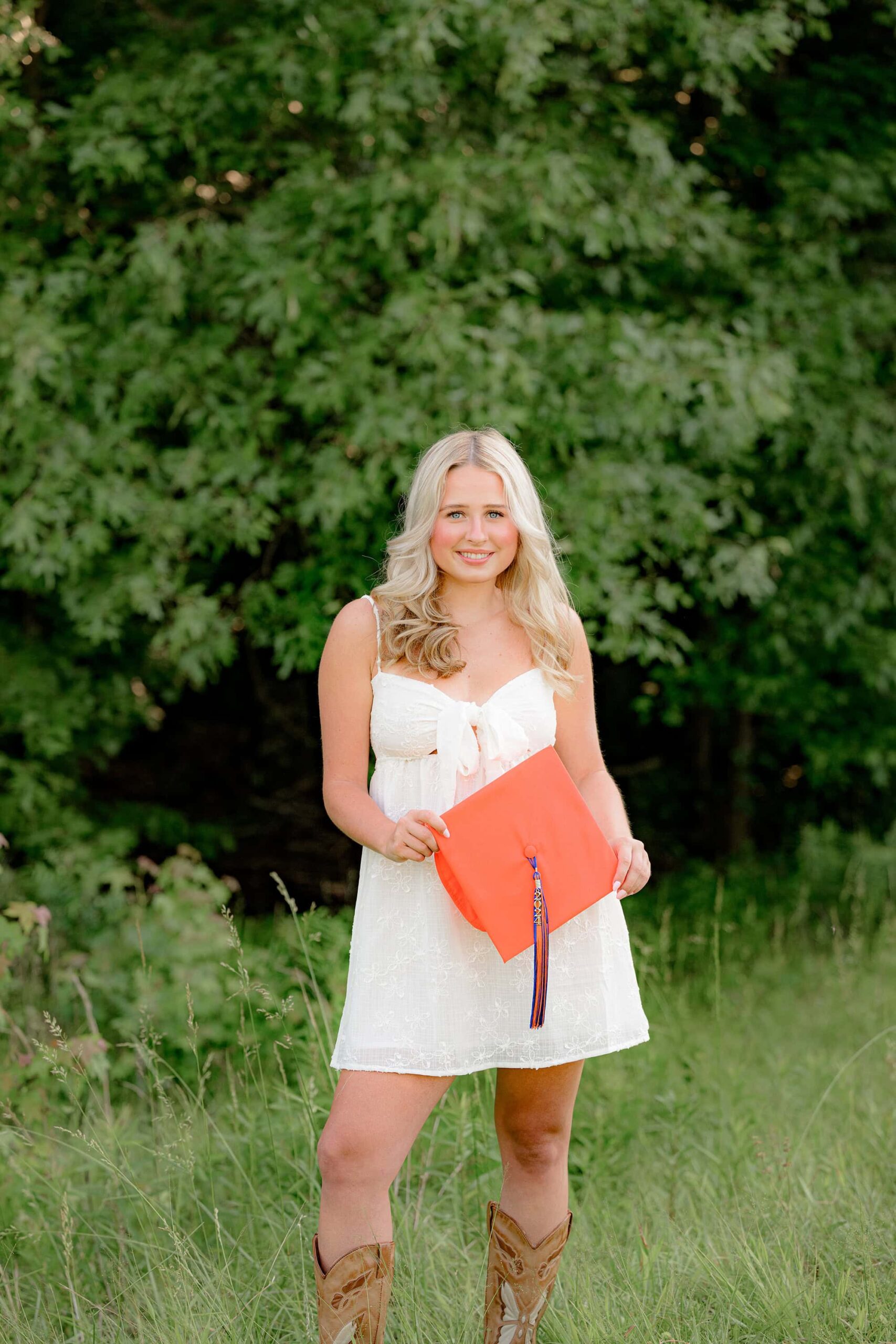 Standing in the tall grass, Kait holds her orange Marvin Ridge High School graduation cap at her waist, with the tassel dangling down during her Charlotte, NC senior photo session