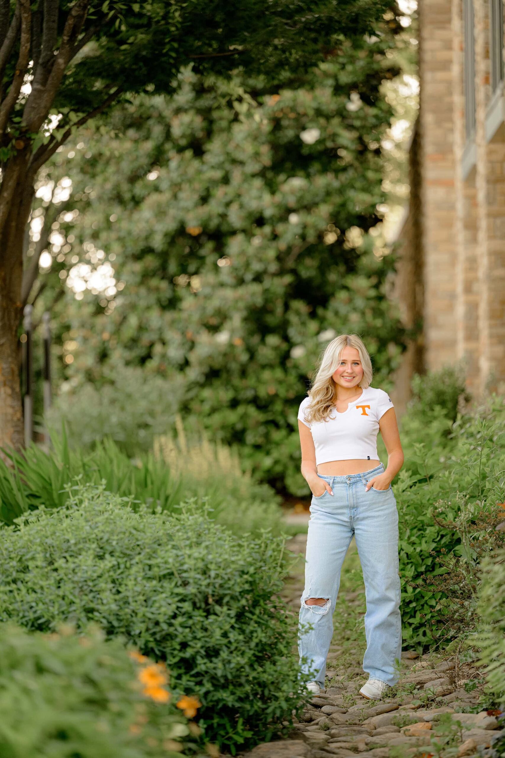 Kait, a Marvin Ridge High School senior, poses for a spring senior photo in a full-body shot. She stands confidently with her hands tucked into the pockets of her jeans, wearing a white crop top featuring the University of Tennessee logo.