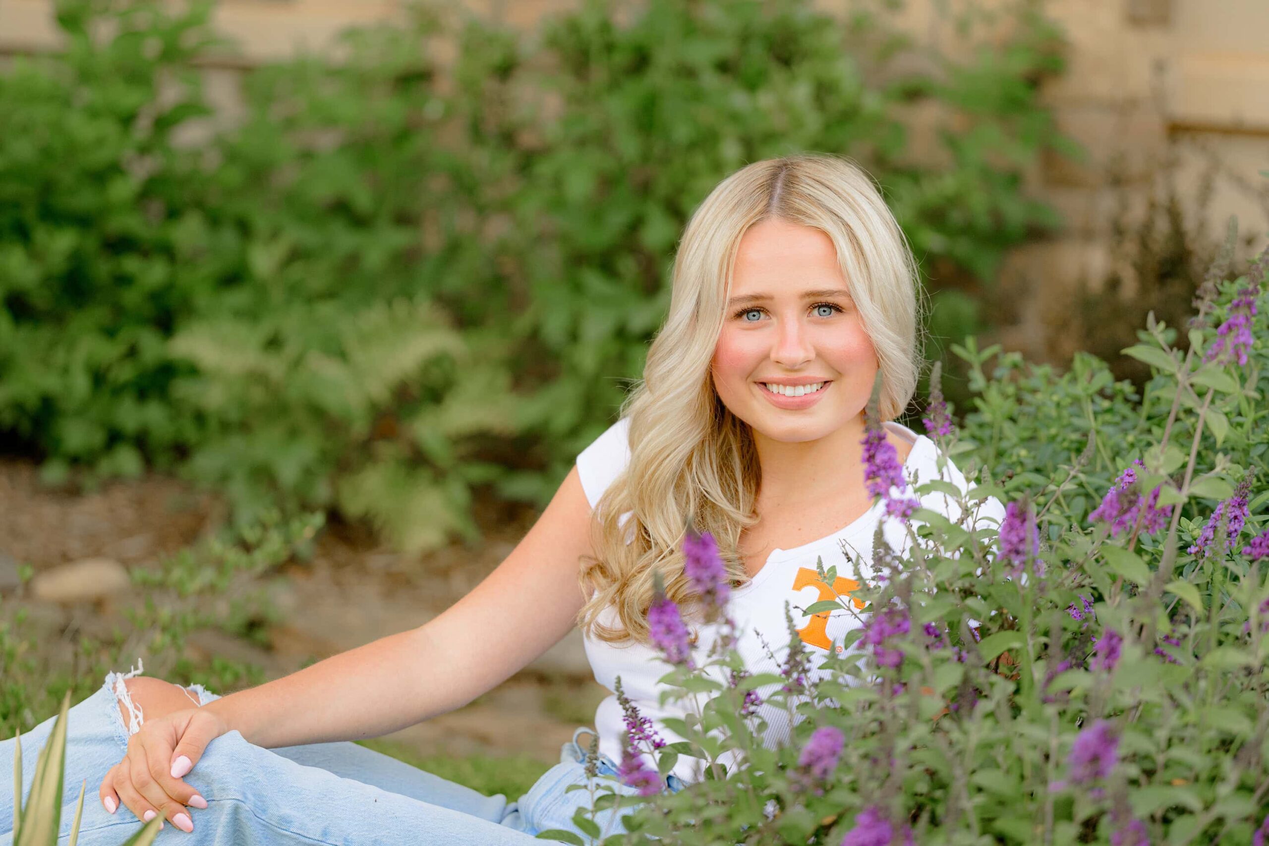 Kait, a Marvin Ridge High School senior, sits in a flower bed surrounded by vibrant purple blooms. She smiles softly, embracing the beauty of spring as she celebrates her senior year.