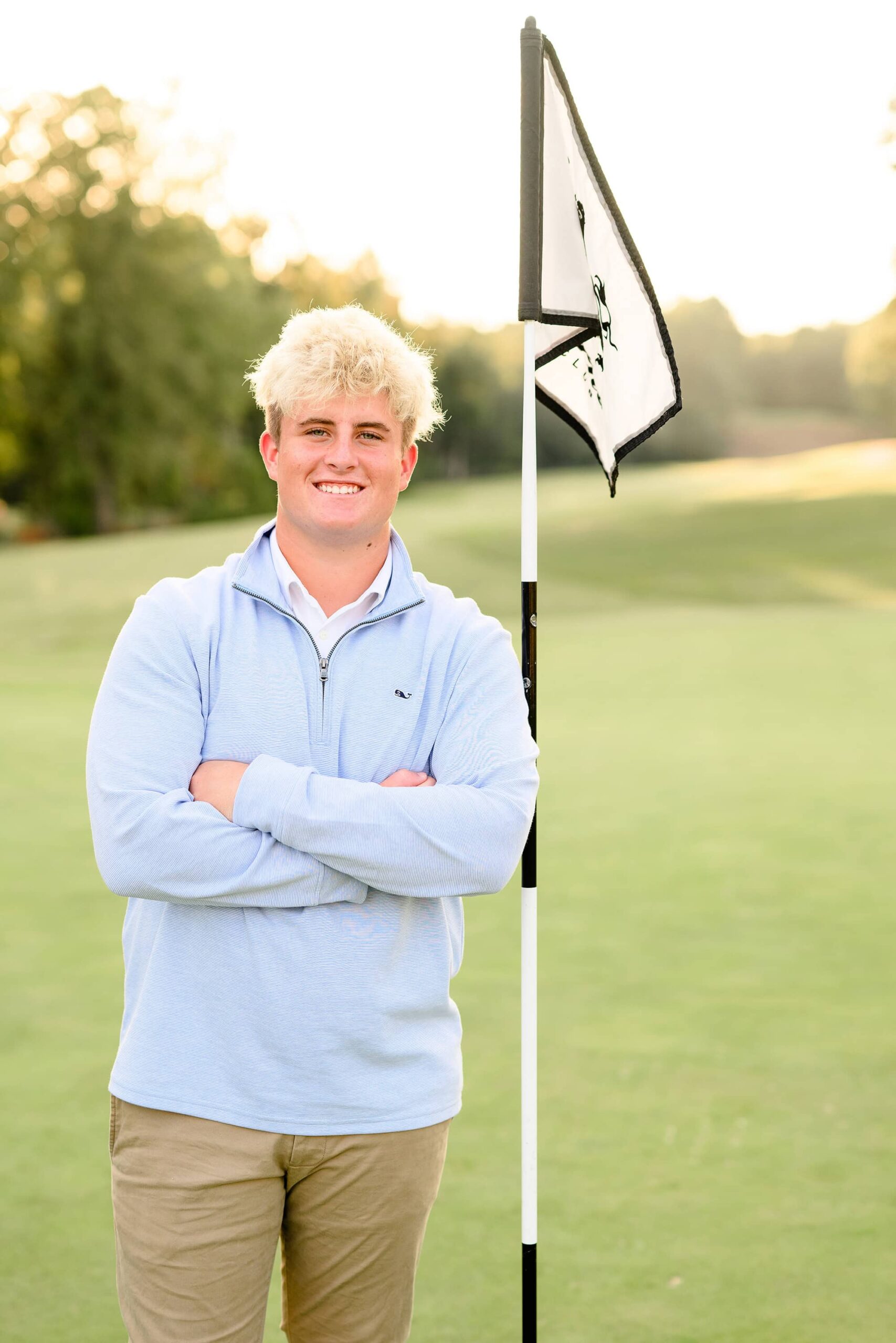 A Marvin Ridge high school senior golfer with curly blonde hair and a light blue quarter-zip sweater stands on a green golf course, smiling with his arms crossed next to a Firethorn golf flag. The sun casts a warm glow in the background.