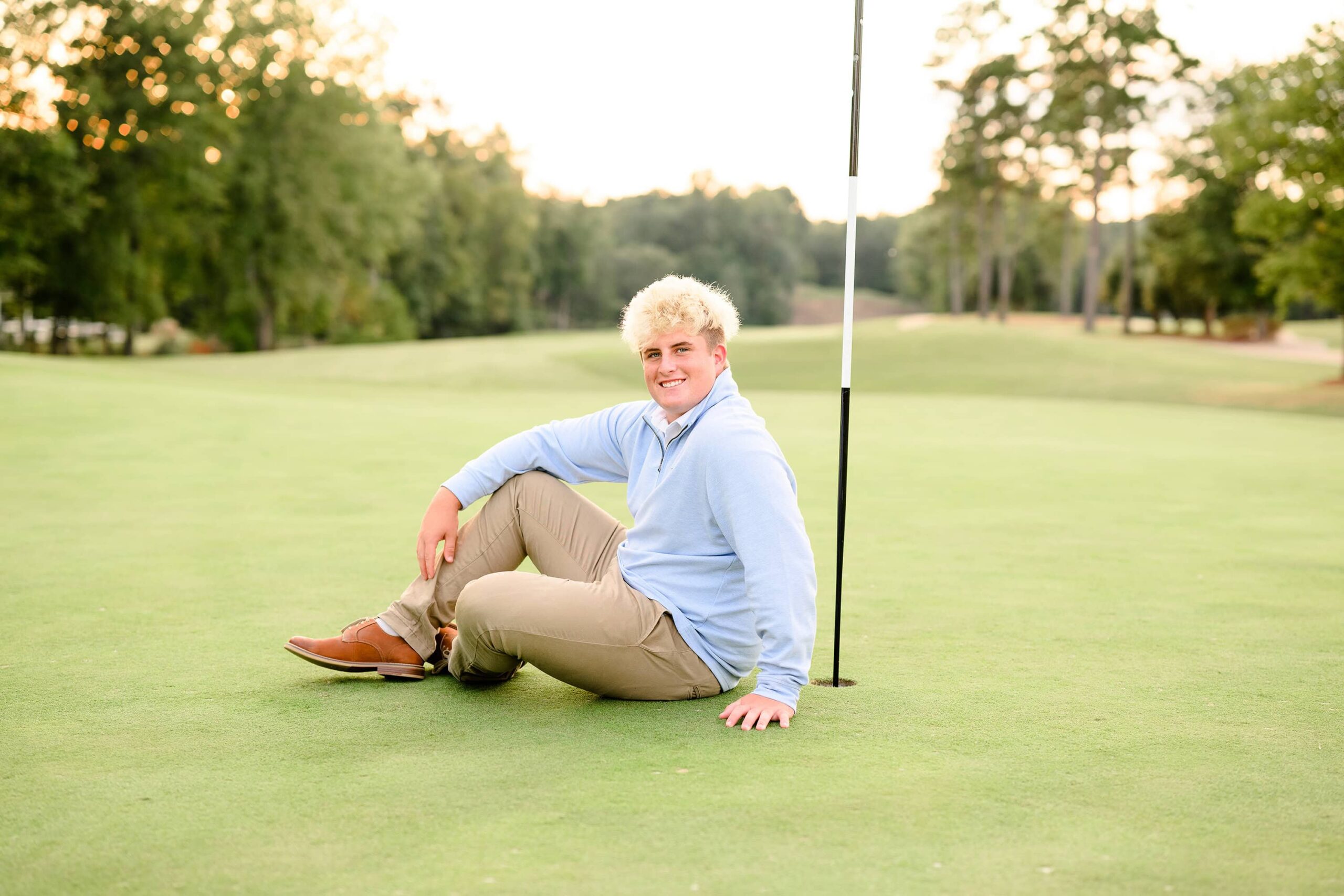 A senior golfer sits on the green, leaning on his hand beside the golf hole. Dressed in a light blue sweater and khakis, he smiles warmly, capturing the joy of his high school senior sports golf journey.