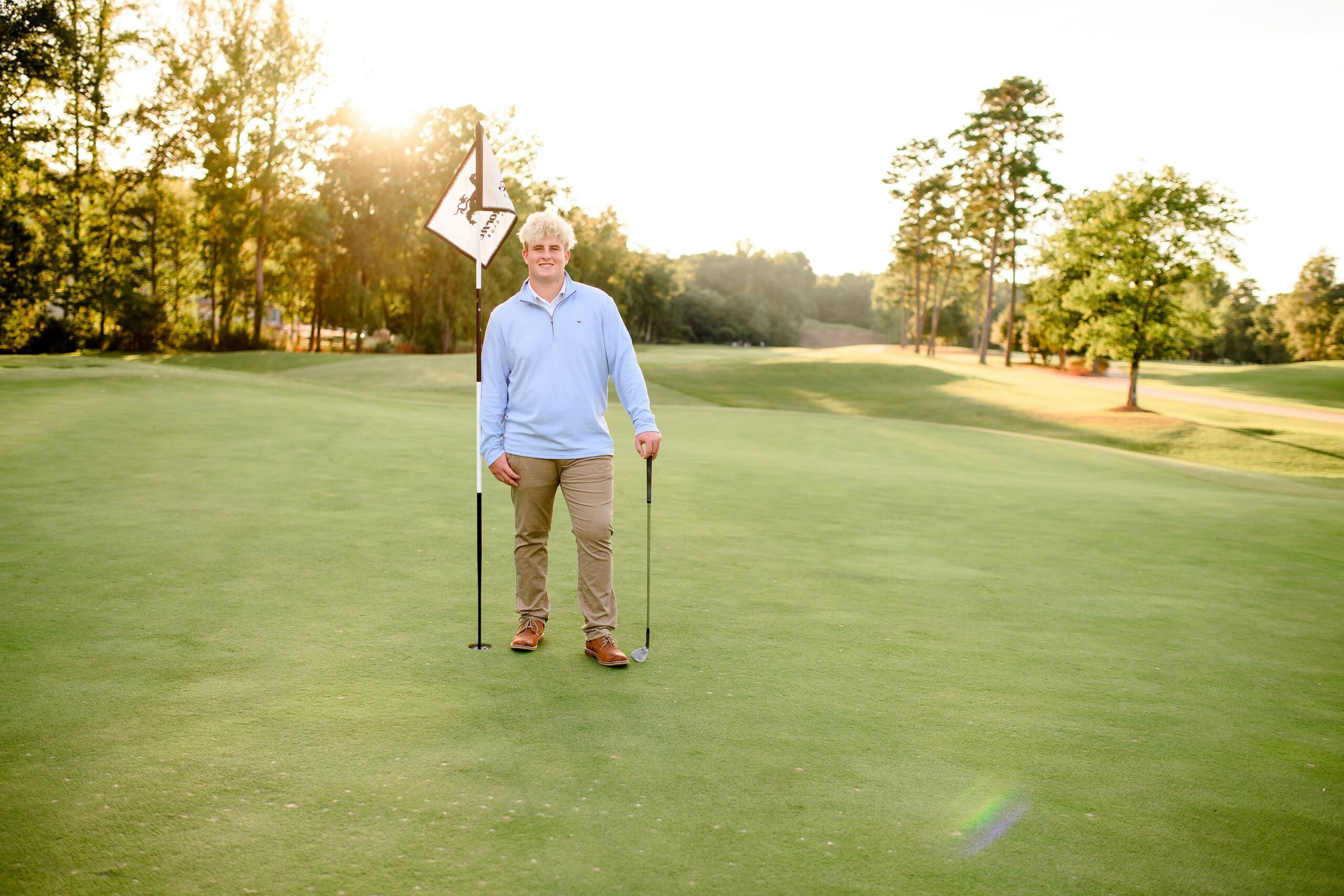 A Marvin Ridge High School senior boy smiles warmly as he holds his golf club in one hand and leans slightly against a golf pole during his senior sports session at Firethorn Country Club.