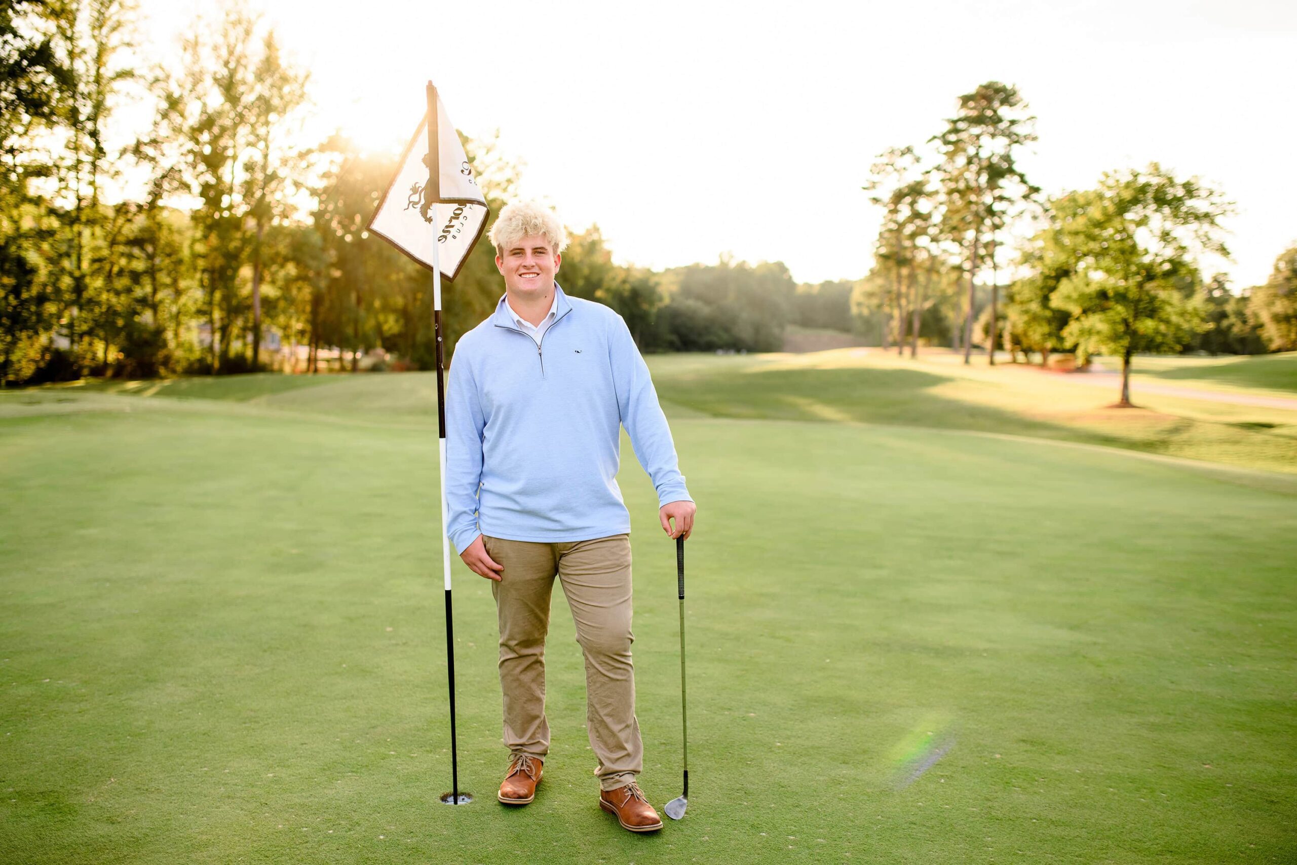 A senior golfer stands confidently next to a hole at Firethorn Country Club, holding a club in one hand and resting the other on the flagstick. The lush golf course and golden-hour sunlight create a serene and picturesque scene.