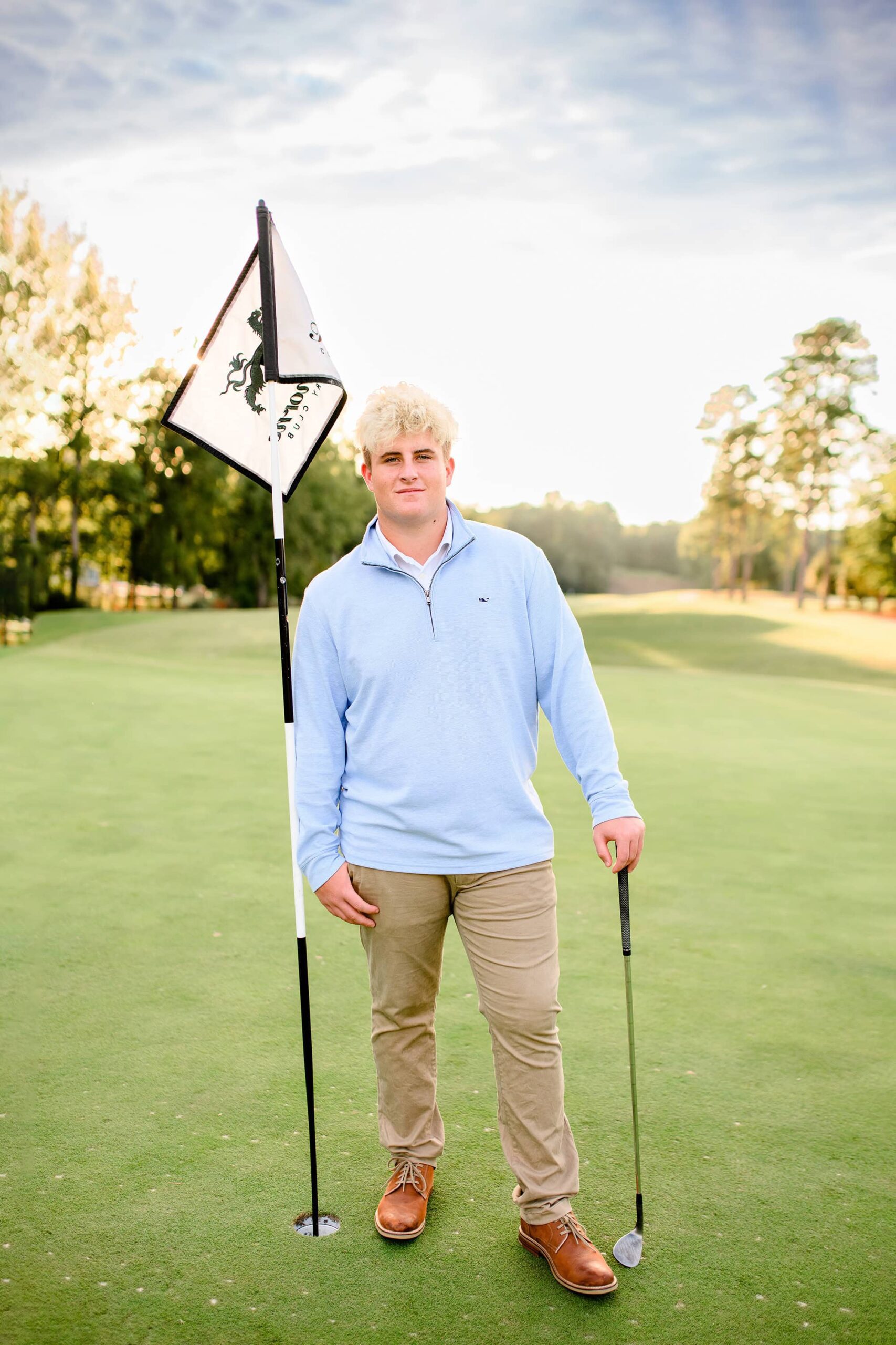 A high school senior golfer stands next to the flagstick at Firethorn Country Club, his thumb casually hooked in his pocket while holding his golf club beside him, its head resting on the green. The golden sunlight highlights the peaceful atmosphere of the course, marking the end of his high school golf journey.