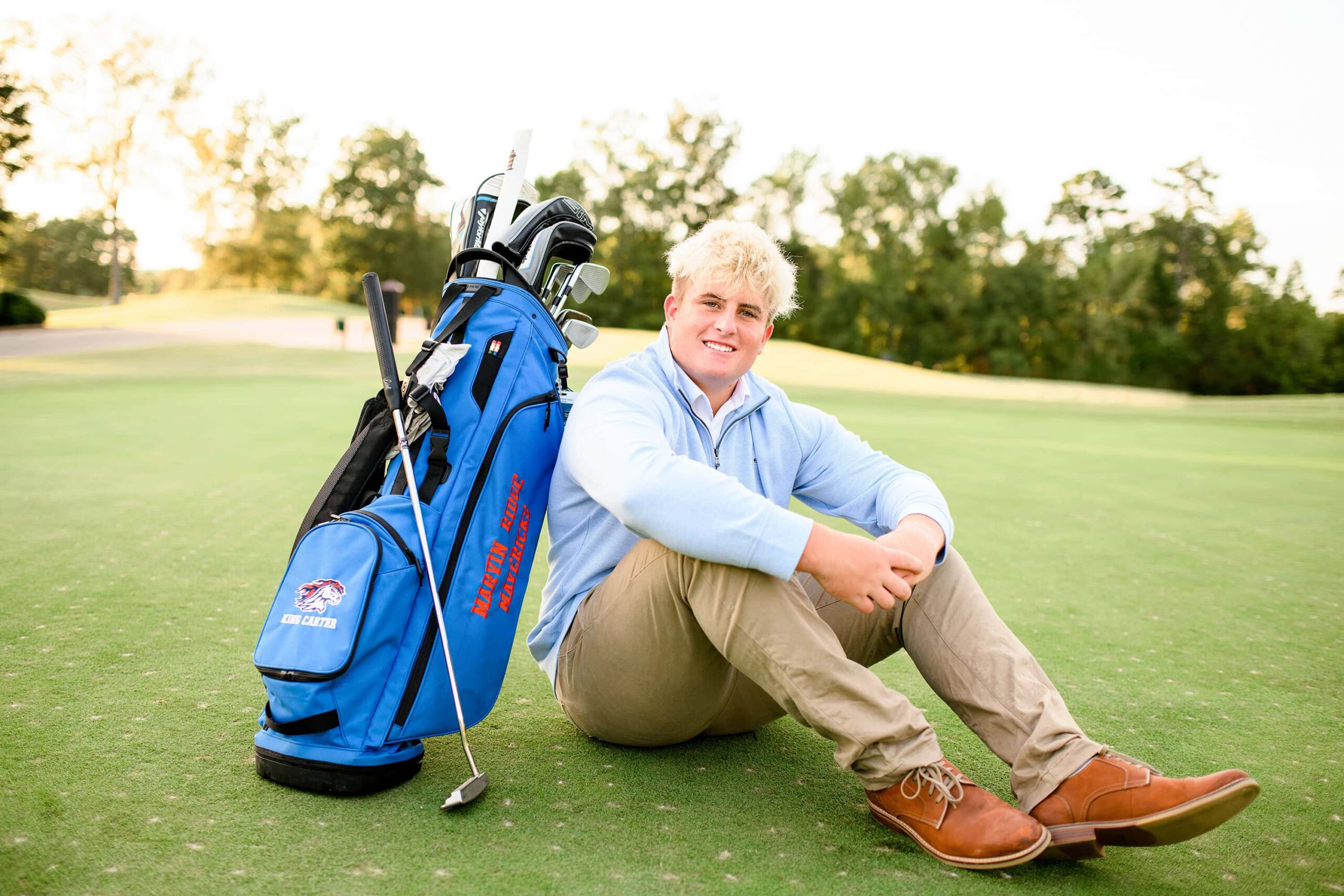 A senior golfer sits on the green, leaning against his blue Marvin Ridge High School golf bag with clubs inside. Dressed in a light blue sweater and khakis, he smiles warmly, capturing the essence of his high school senior sports golf journey.