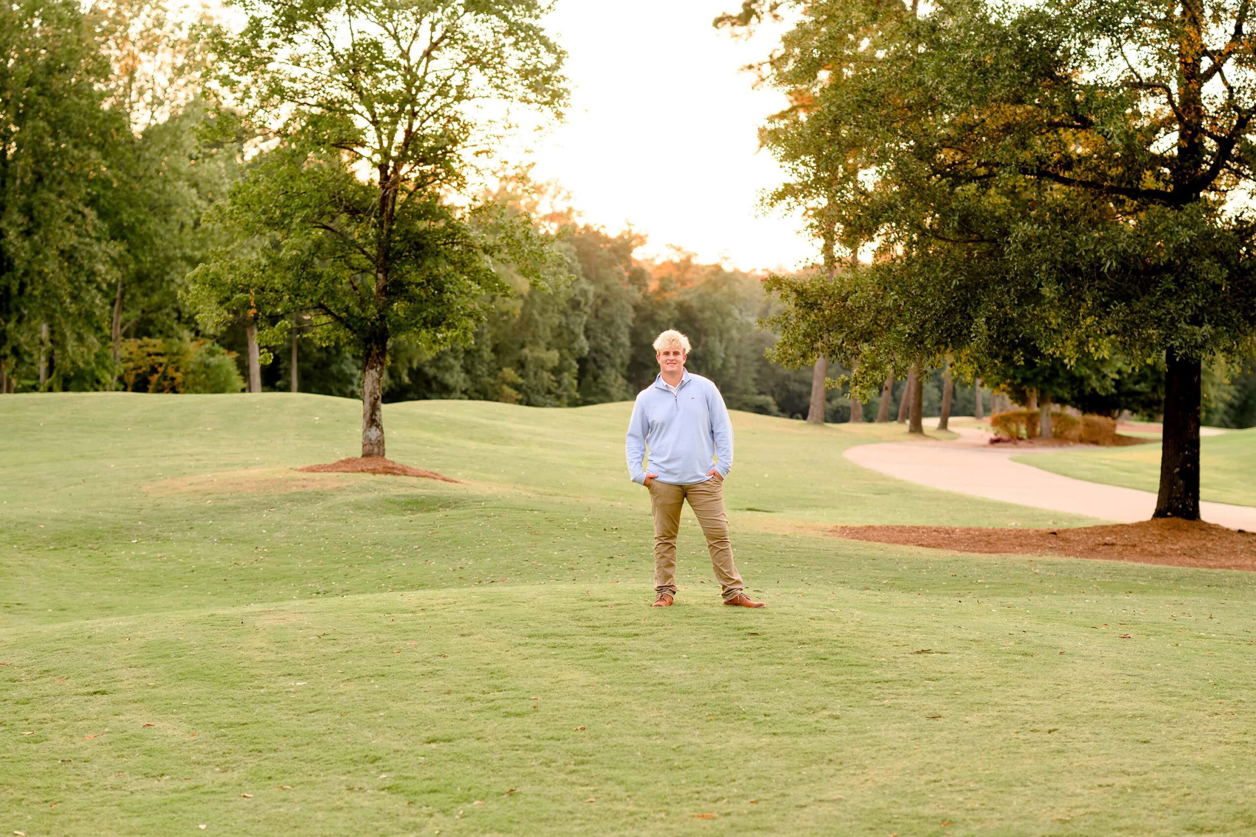 As the sun sets behind him, a senior golfer stands on the course with one hand in his pocket, toward the camera as the sun sets behind him.