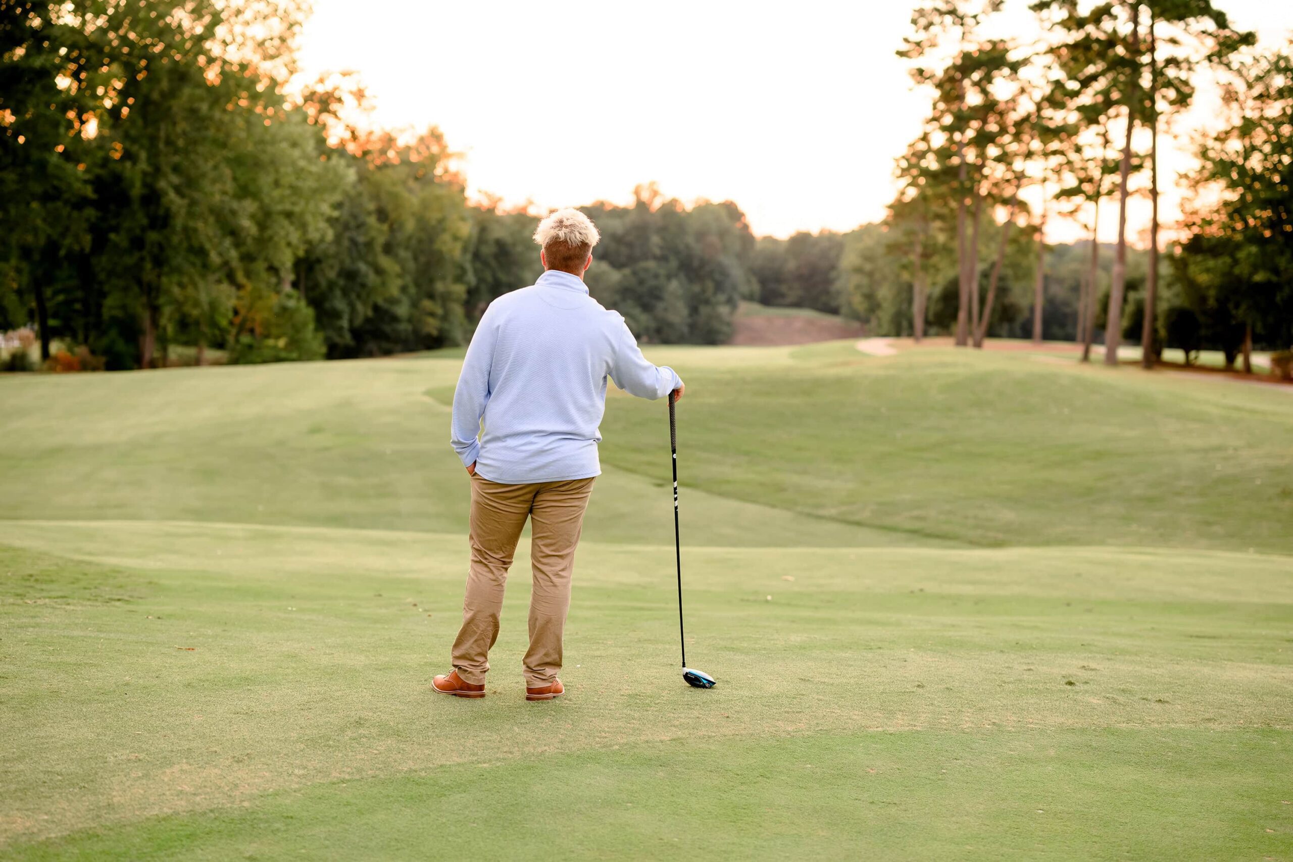 As the sun sets behind him, a senior golfer stands on the course with one hand in his pocket, gazing into the distance. His back is turned to the camera, silhouetted against the golden glow of the evening light, capturing a reflective and timeless moment.
