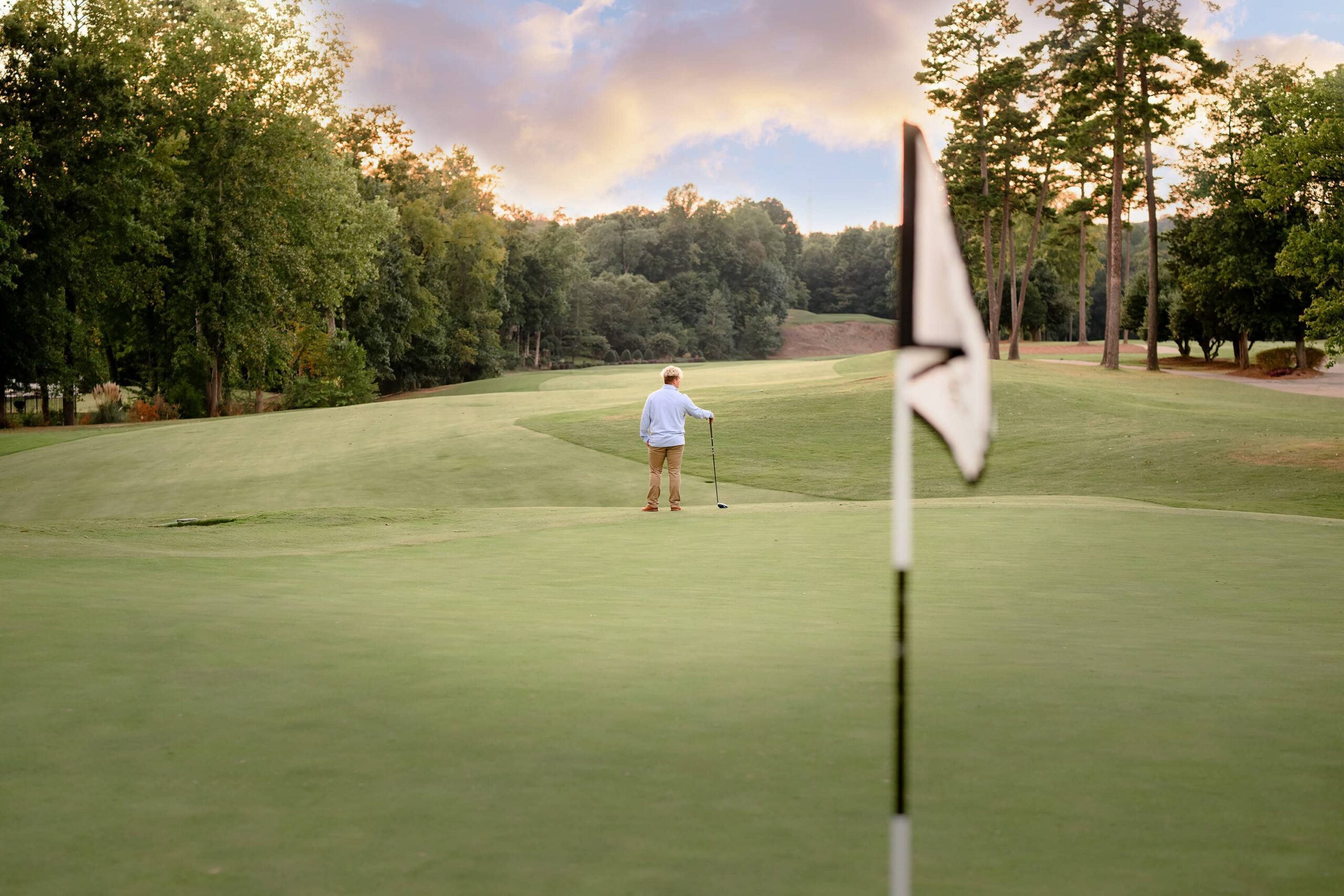 A senior golfer stands off in the distance on the golf course, one hand in his pocket and the other holding a club. In the foreground, a golf flag flutters, creating a dynamic composition with the lush green fairway stretching behind him.