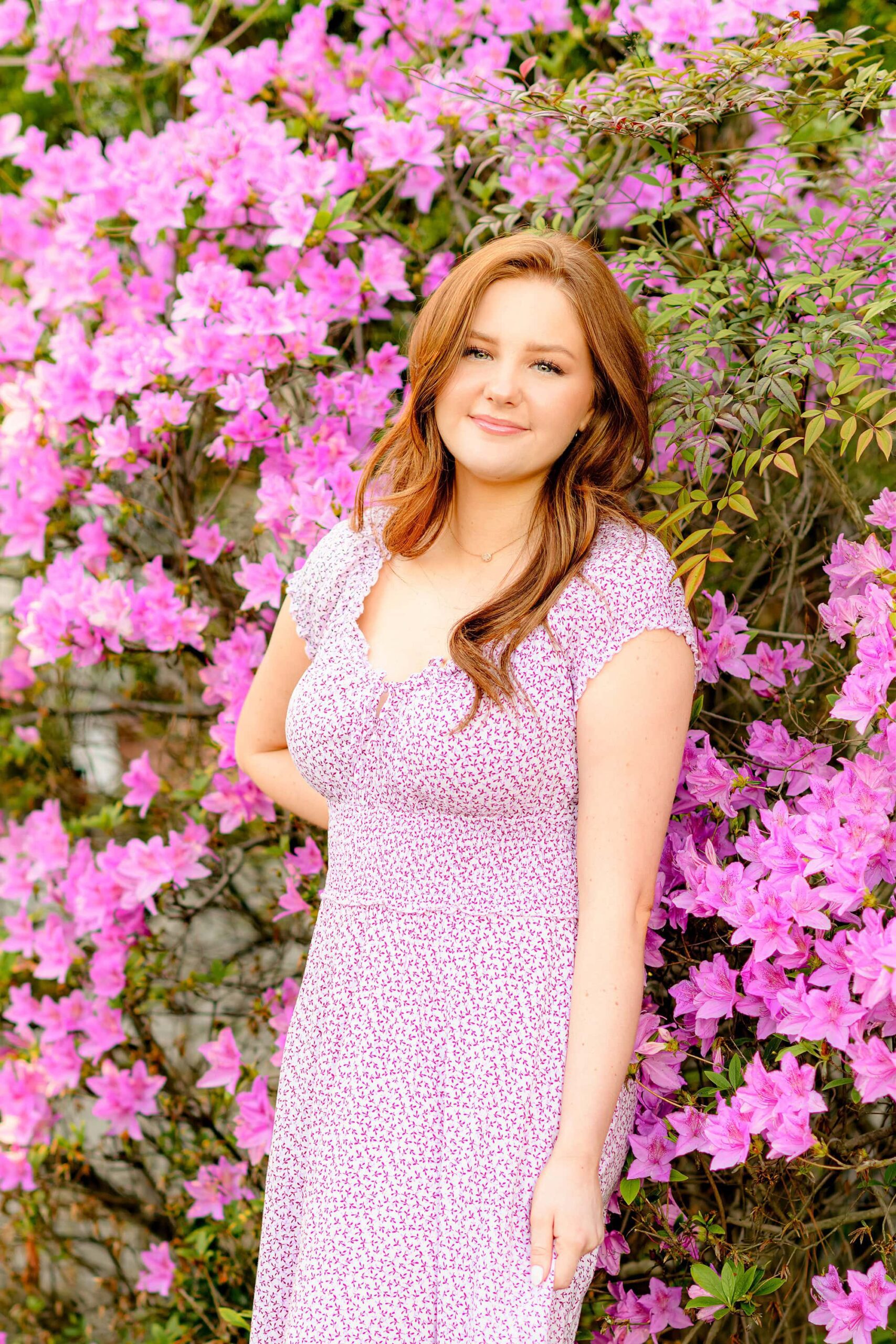 The red-headed senior holds the fabric of her dress while smirking playfully, surrounded by blooming azaleas, creating a lively and joyful vibe.