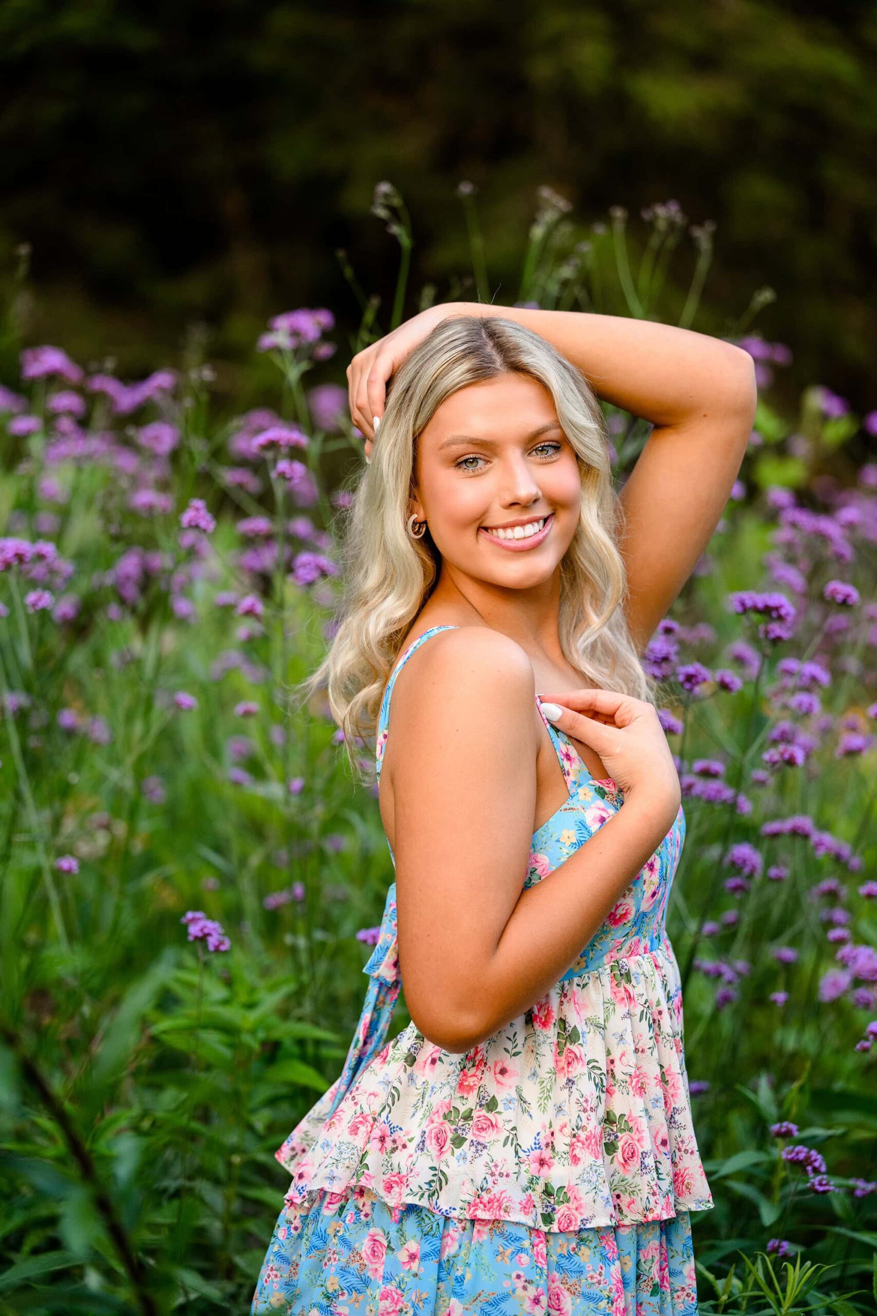 A blonde senior in a tiered flower dress stands in a vibrant flower garden in Waxhaw, NC, surrounded by purple verbena and white flowers. She poses with her arms gracefully around her head, capturing a serene moment in her senior photos in flowers.