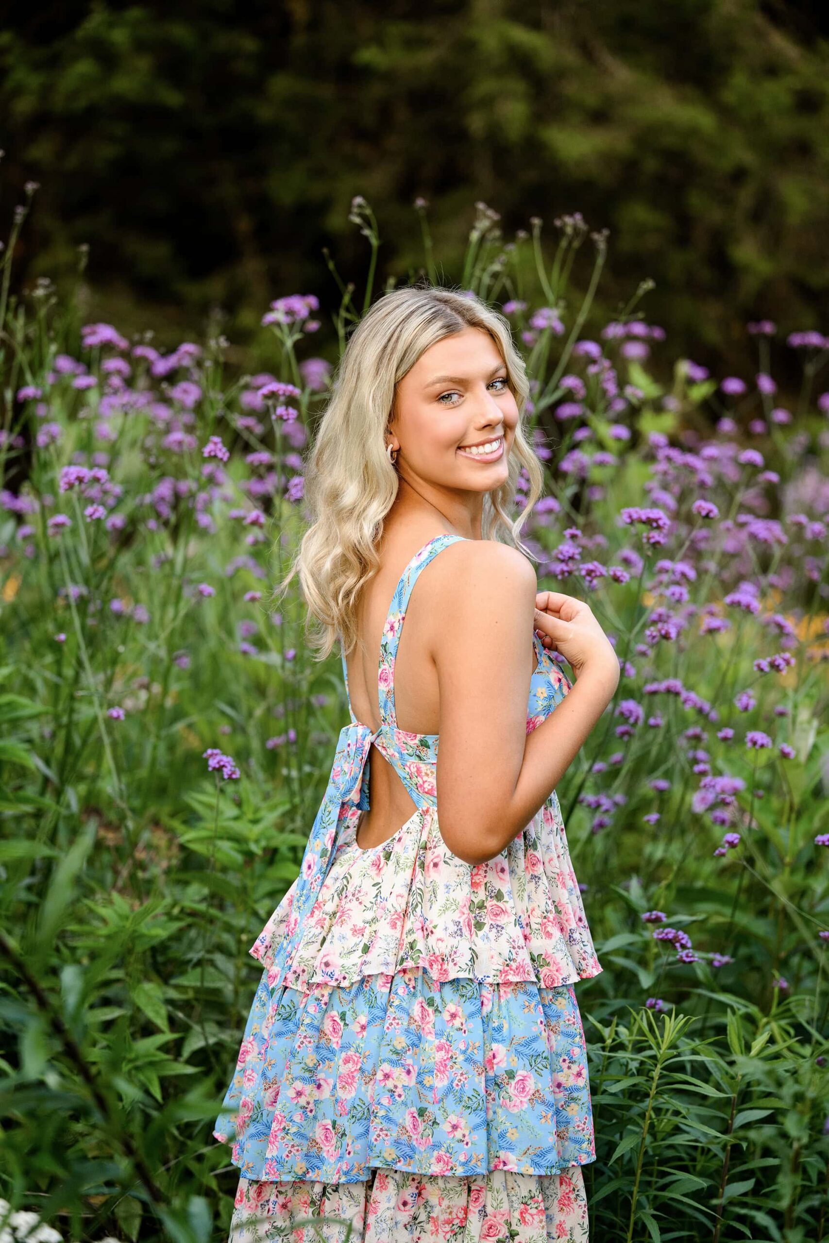 The blonde Cuthbbertson High School senior looks back over her shoulder, showcasing the decorative back of her tiered flower dress. The purple verbena and white flowers create a stunning backdrop, completing her graceful pose in the flower garden.