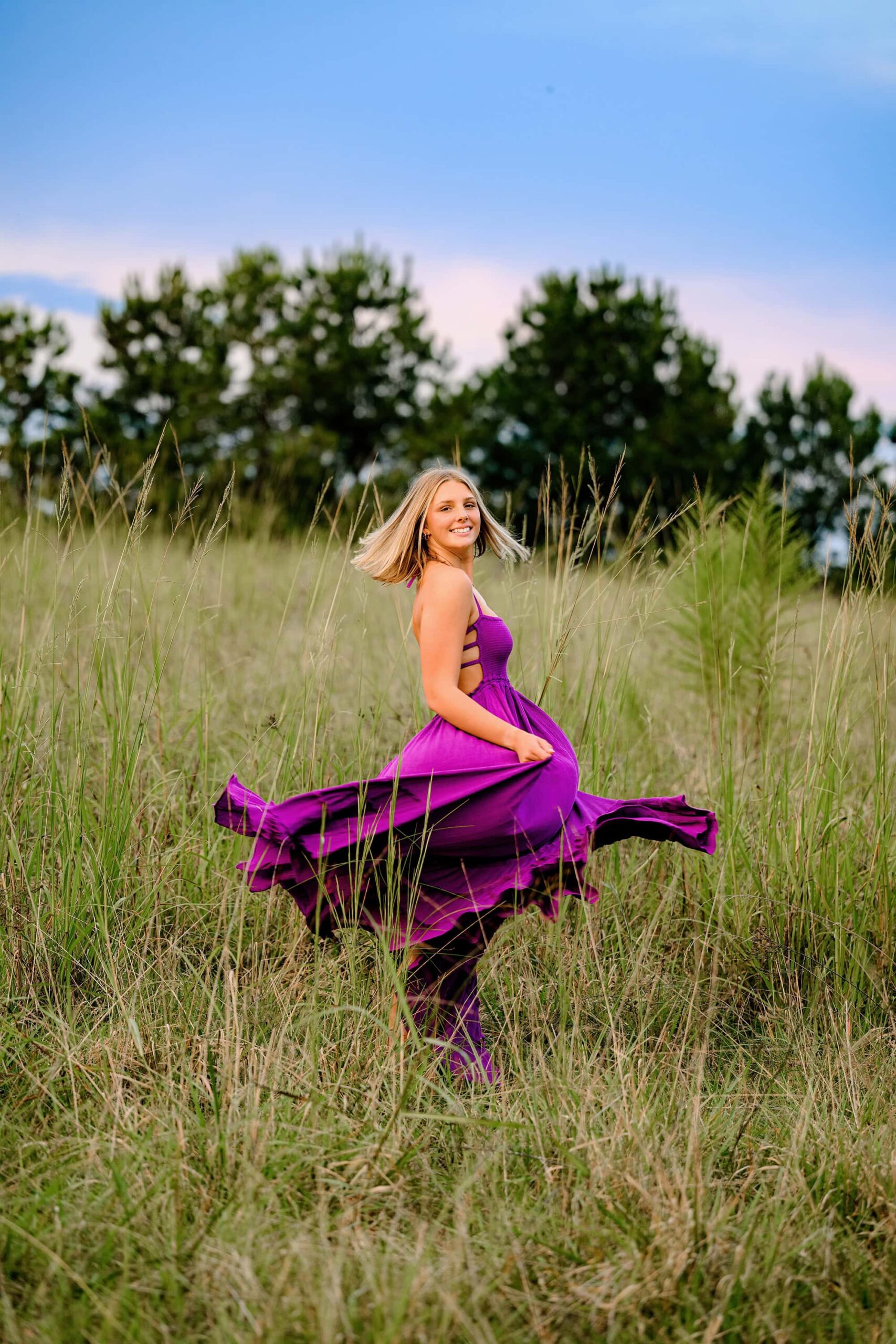The blonde senior twirls gracefully in an open field, her long purple dress flowing around her. The movement captures a joyful and carefree spirit as she enjoys the natural beauty of the surroundings.
