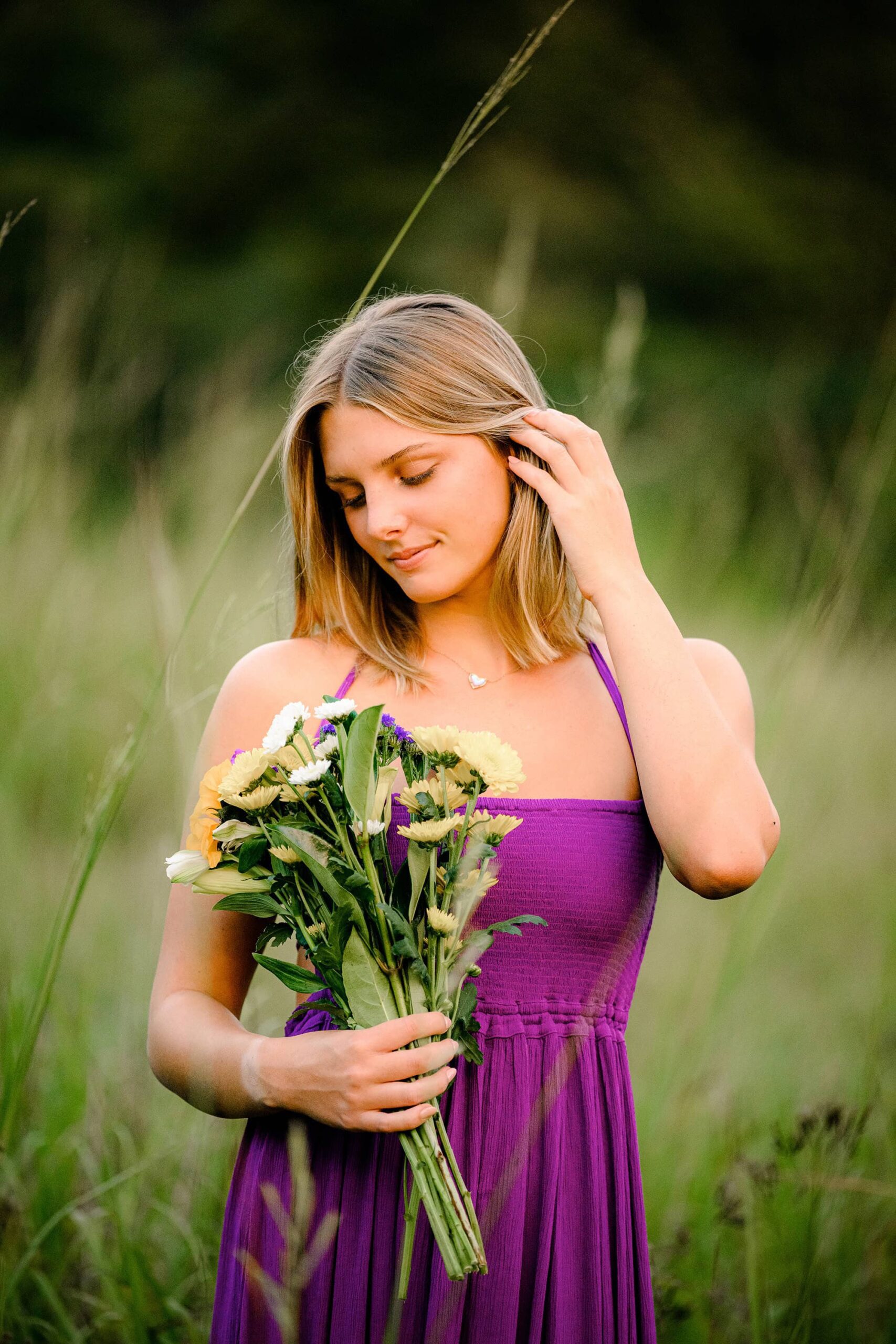 A short-haired blonde senior from Union County, wearing a long purple dress, looks down at a bouquet of flowers, creating a soft, serene moment in an open field.