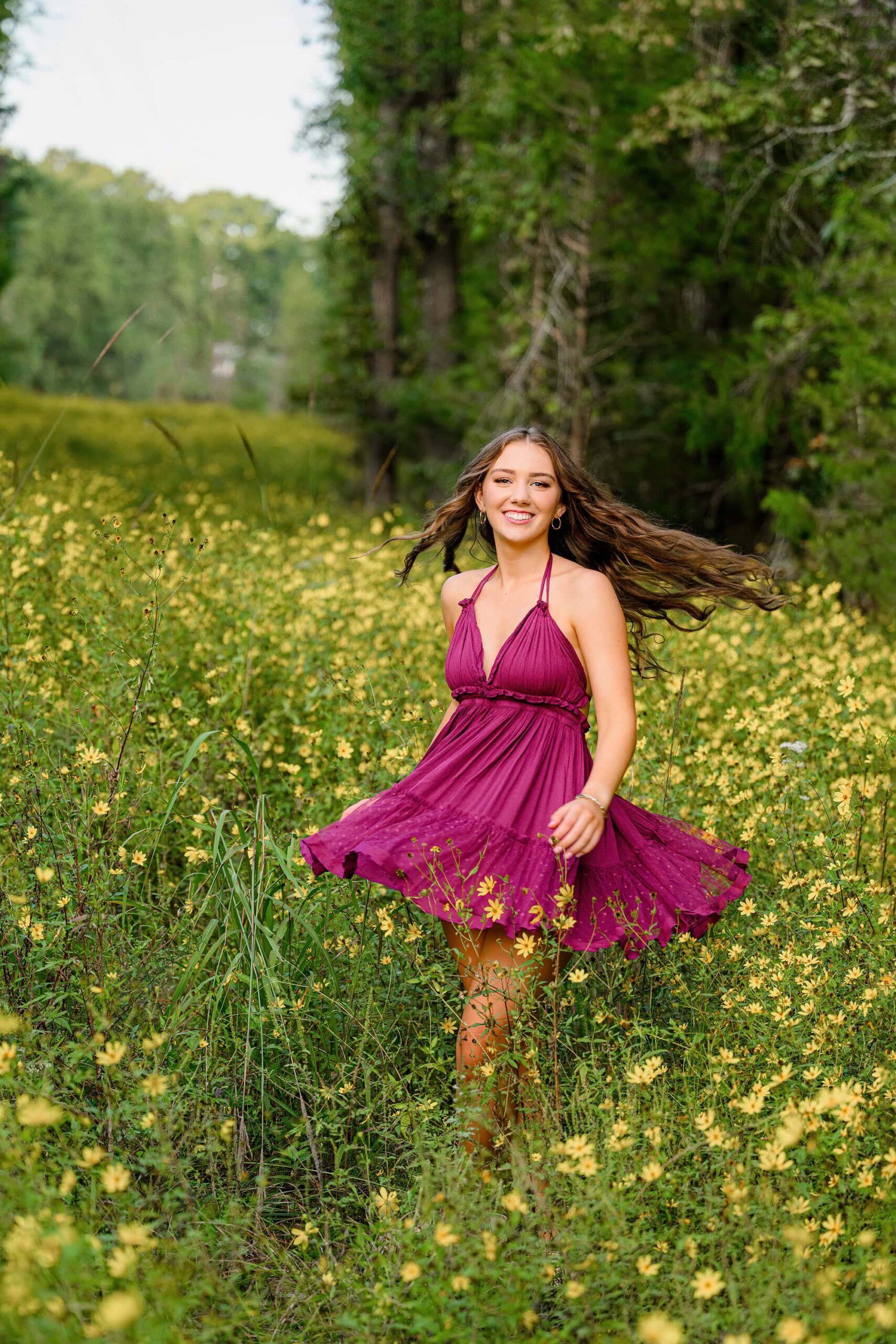 A long-haired senior graduate in a purple dress twirls in a field of fall wildflowers, her dress and hair flowing out wide. The vibrant flowers surround her, capturing a joyful and free-spirited moment.