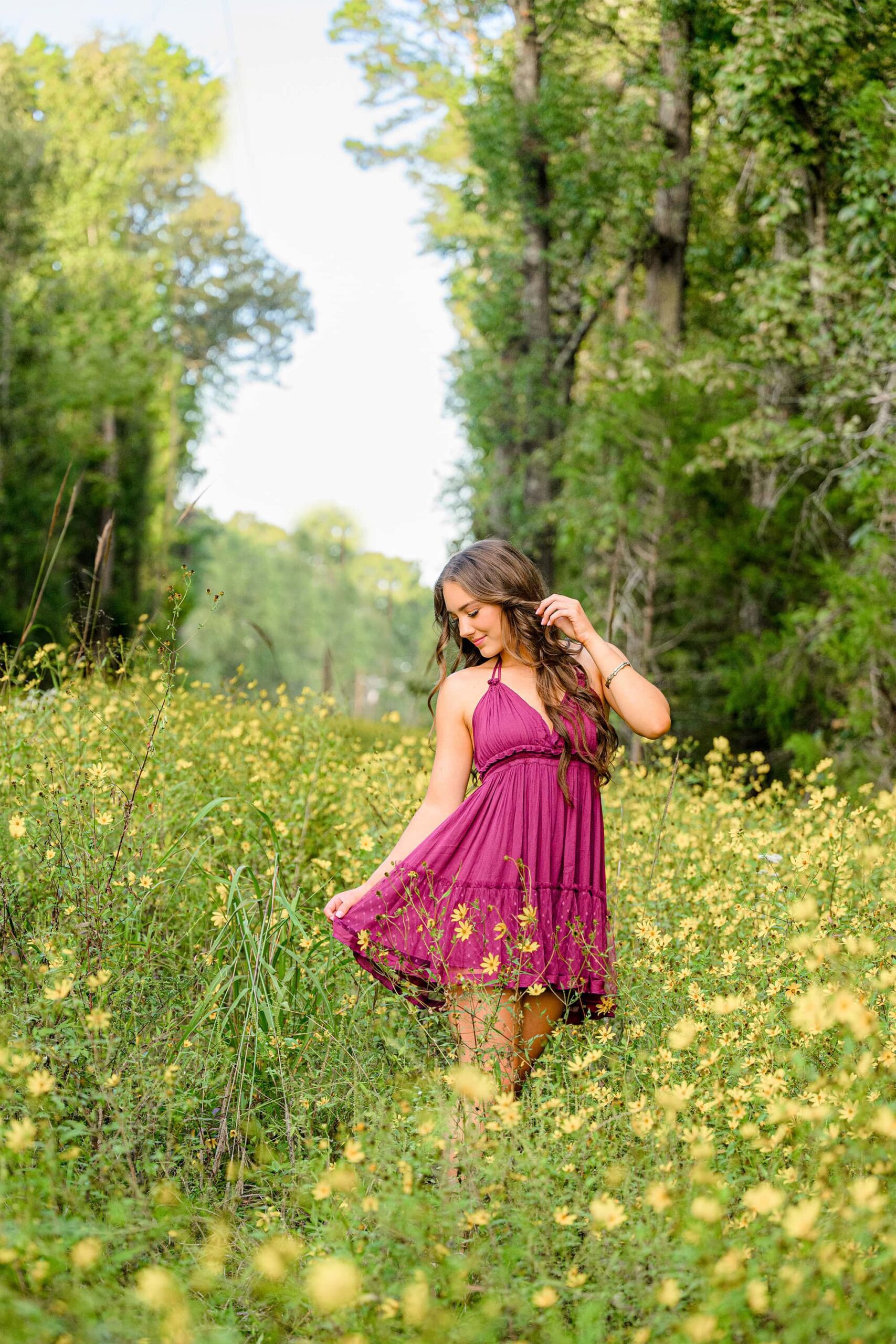 The senior graduate looks down at her dress while tucking her hair behind her ear, standing among the fall wildflowers. Her thoughtful expression and delicate movement add a quiet, reflective charm to the scene.