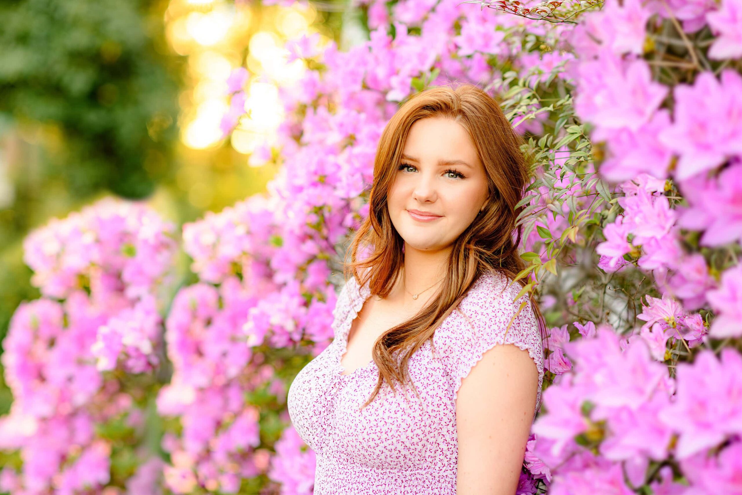 The red-headed senior leans into a lush display of purple azaleas, with soft golden light glowing behind her, creating a dreamy and serene atmosphere.