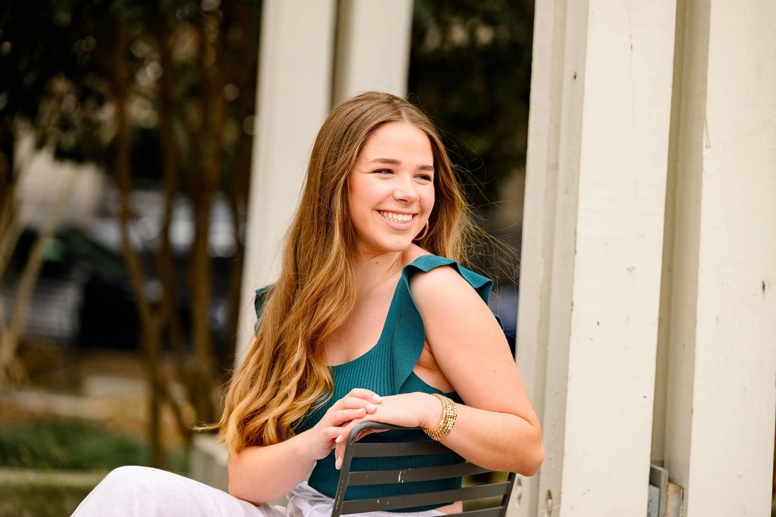 The senior is dressed in a teal short-sleeve sweater and white jeans, sitting in a chair in uptown Charlotte. She looks off-camera, creating a reflective and stylish moment.