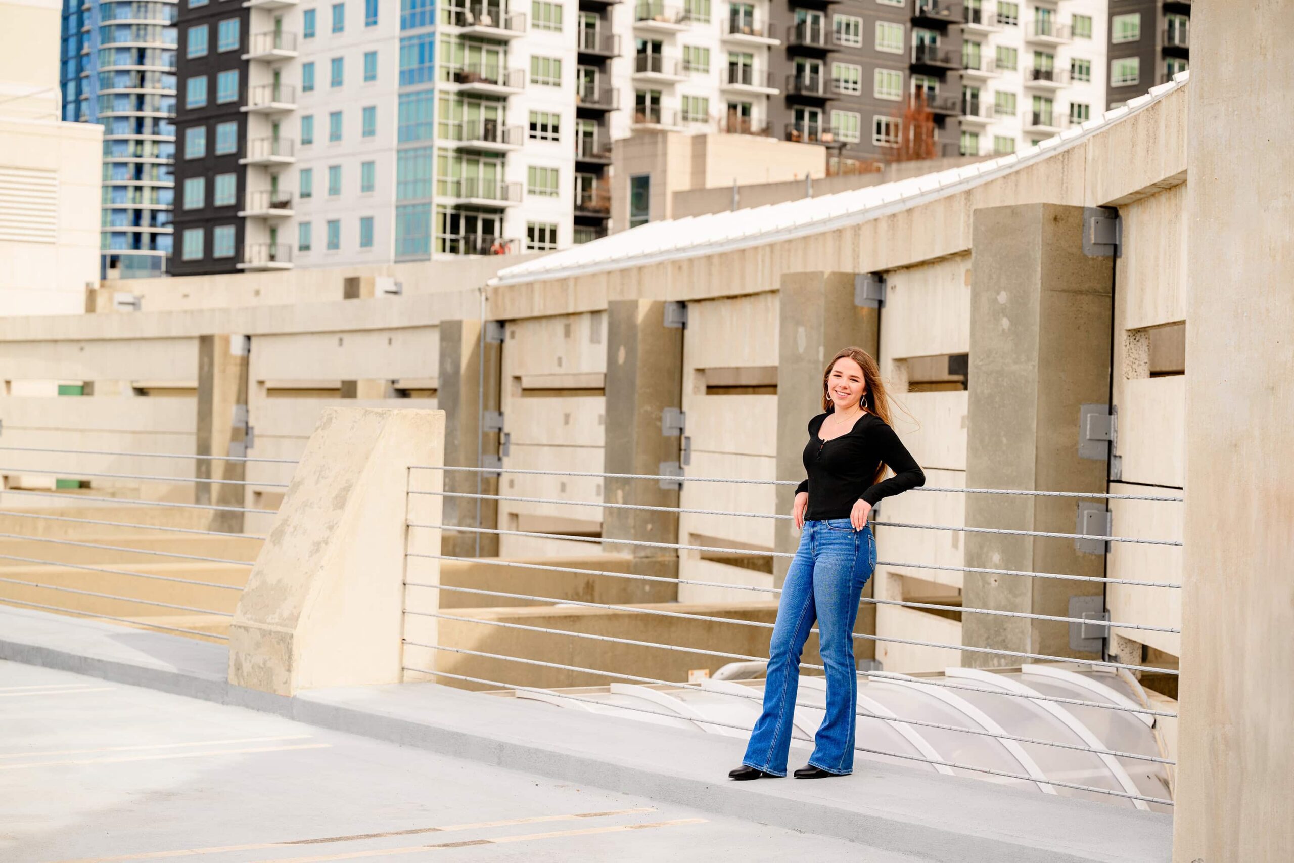 The senior wears jeans and a black long-sleeve shirt, with her hair styled behind her, as she poses confidently in front of the Charlotte buildings. The urban backdrop adds a modern touch to her winter session.