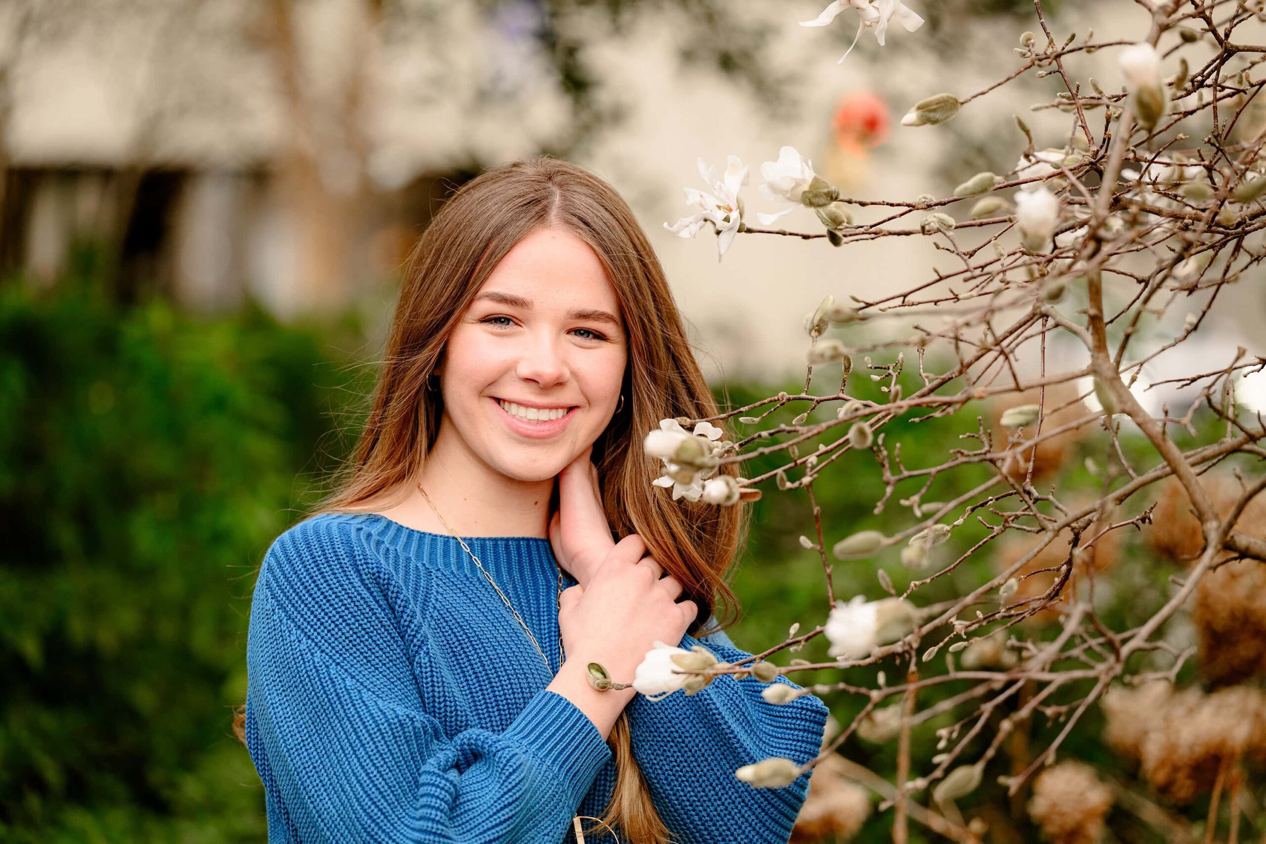 A Waxhaw senior in a blue sweater stands beside a tree with winter flowers in uptown Charlotte. The delicate blooms complement her winter outfit, creating a serene and elegant moment in her senior photos with flowers.